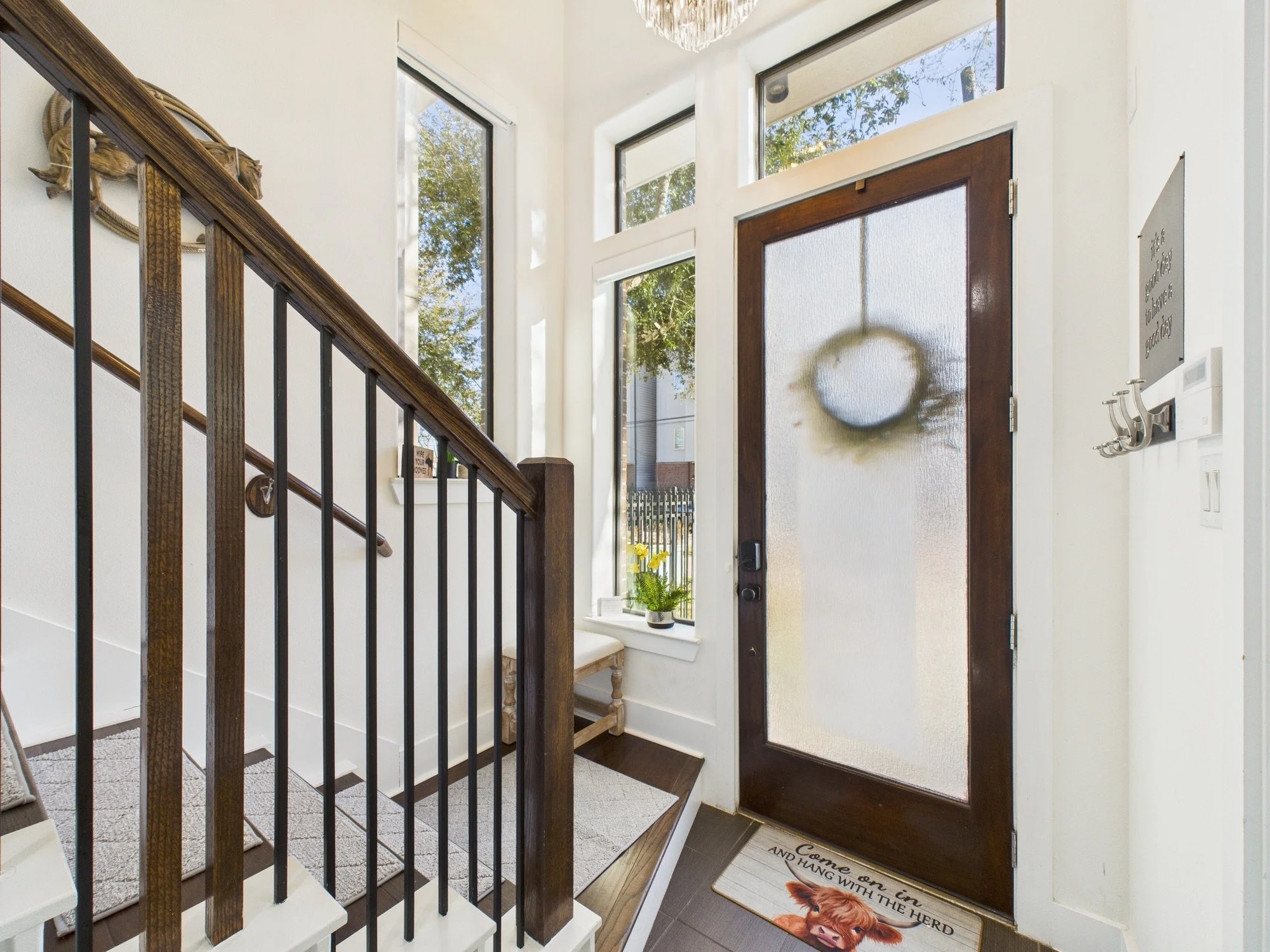 Entryway with wooden front door featuring a frosted glass panel, large floor-to-ceiling windows allowing natural light, a staircase with wooden handrail and metal balusters, a small bench, a potted plant, and a decorative wall sign above hooks.