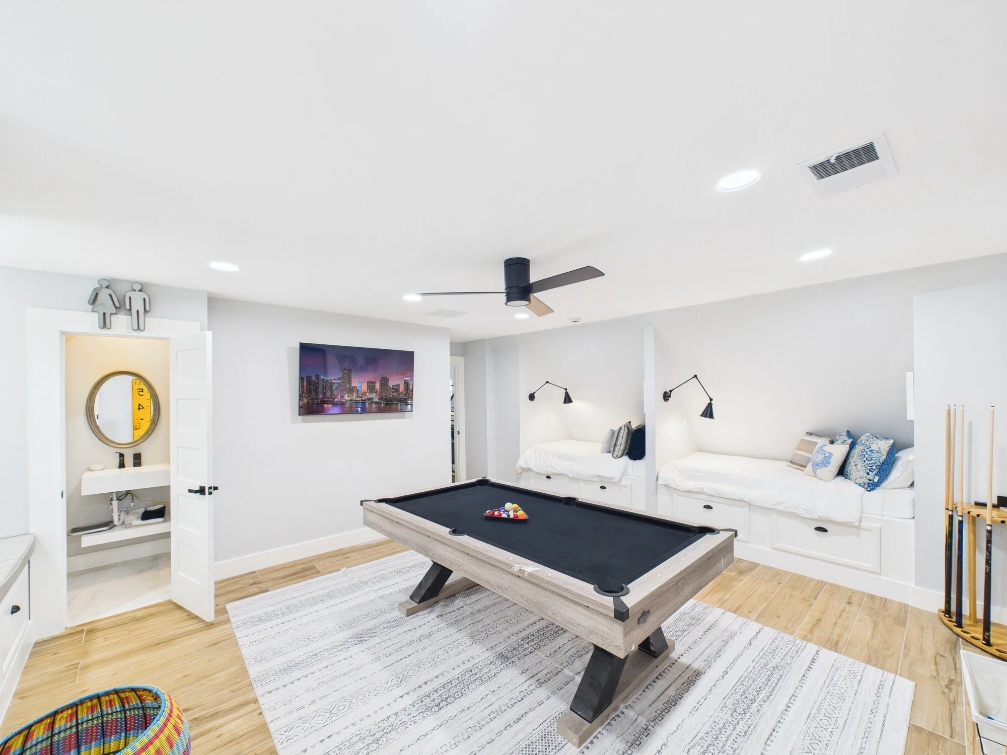 Modern living room with white walls and wood flooring, featuring a pool table with black felt, a large flat-screen TV showing a cityscape, and a black ceiling fan. There are wall-mounted lamps above a white couch, and a door with signage of male and 