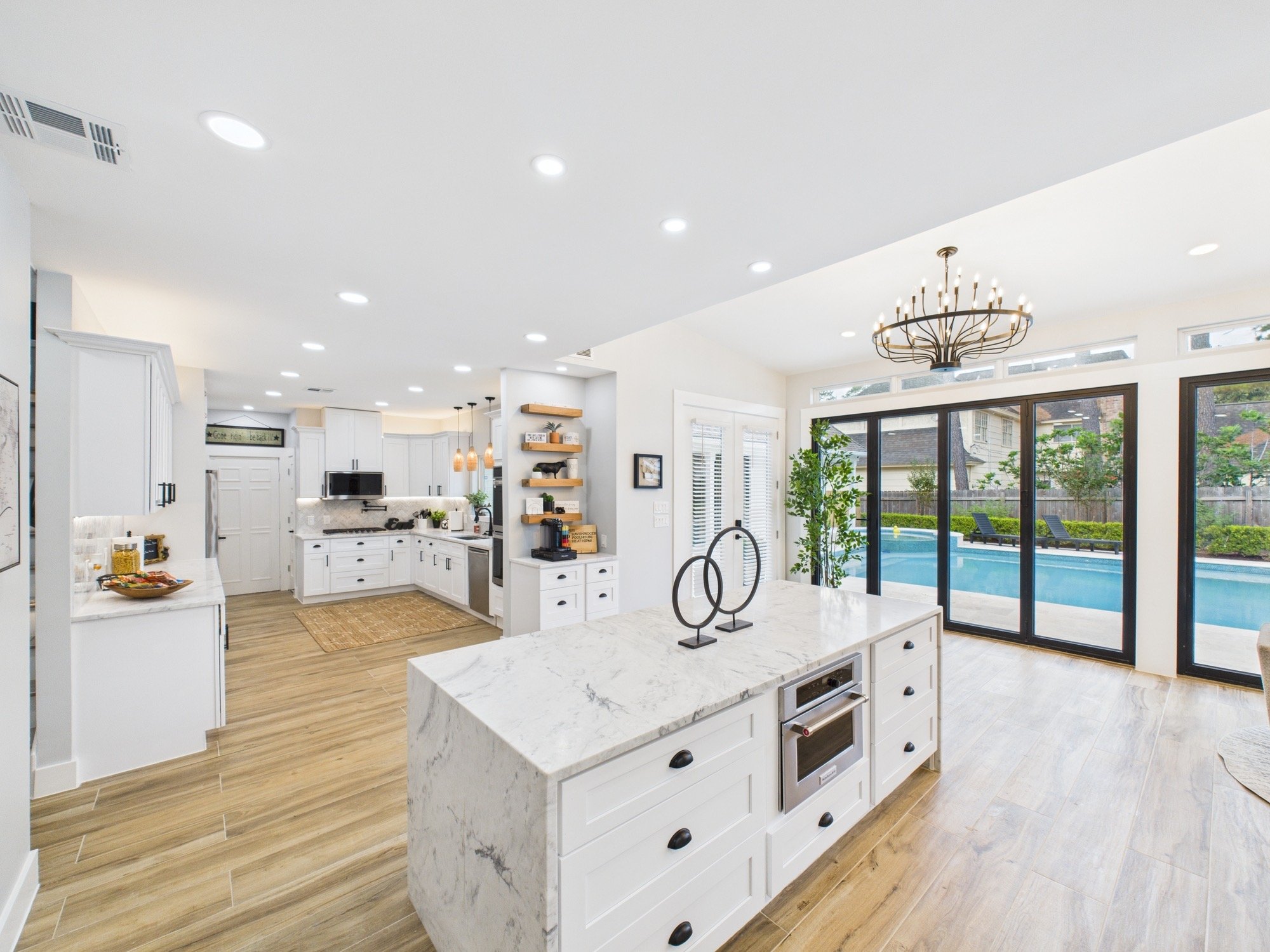 Modern open-concept kitchen and living area with white cabinetry, marble island, hardwood floors, large windows, and outdoor pool view.