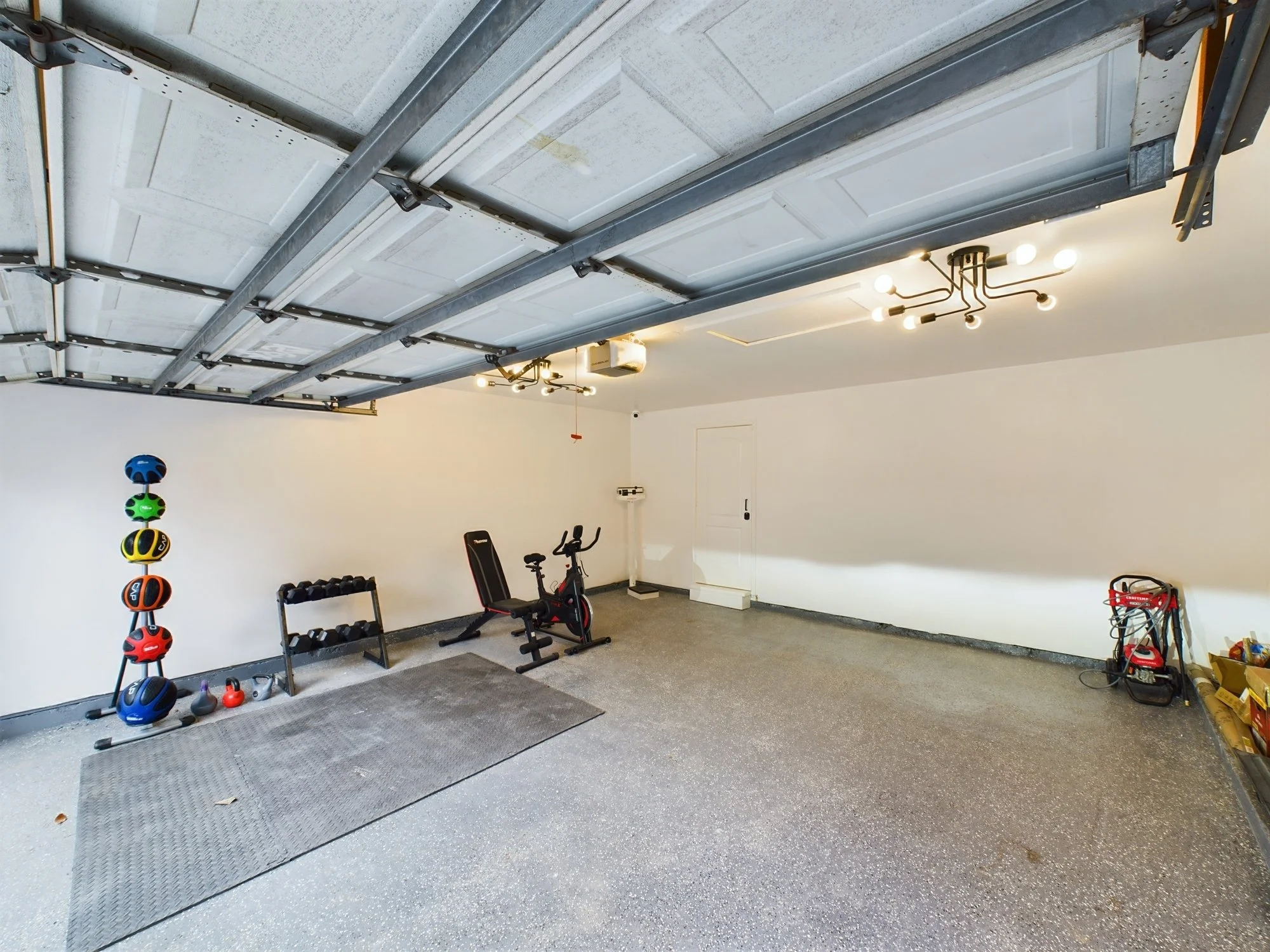 Garage with a partially open garage door, exercise equipment including a stationary bike and dumbbells, colorful helmets on a rack, and some tools and boxes stored in the corner.