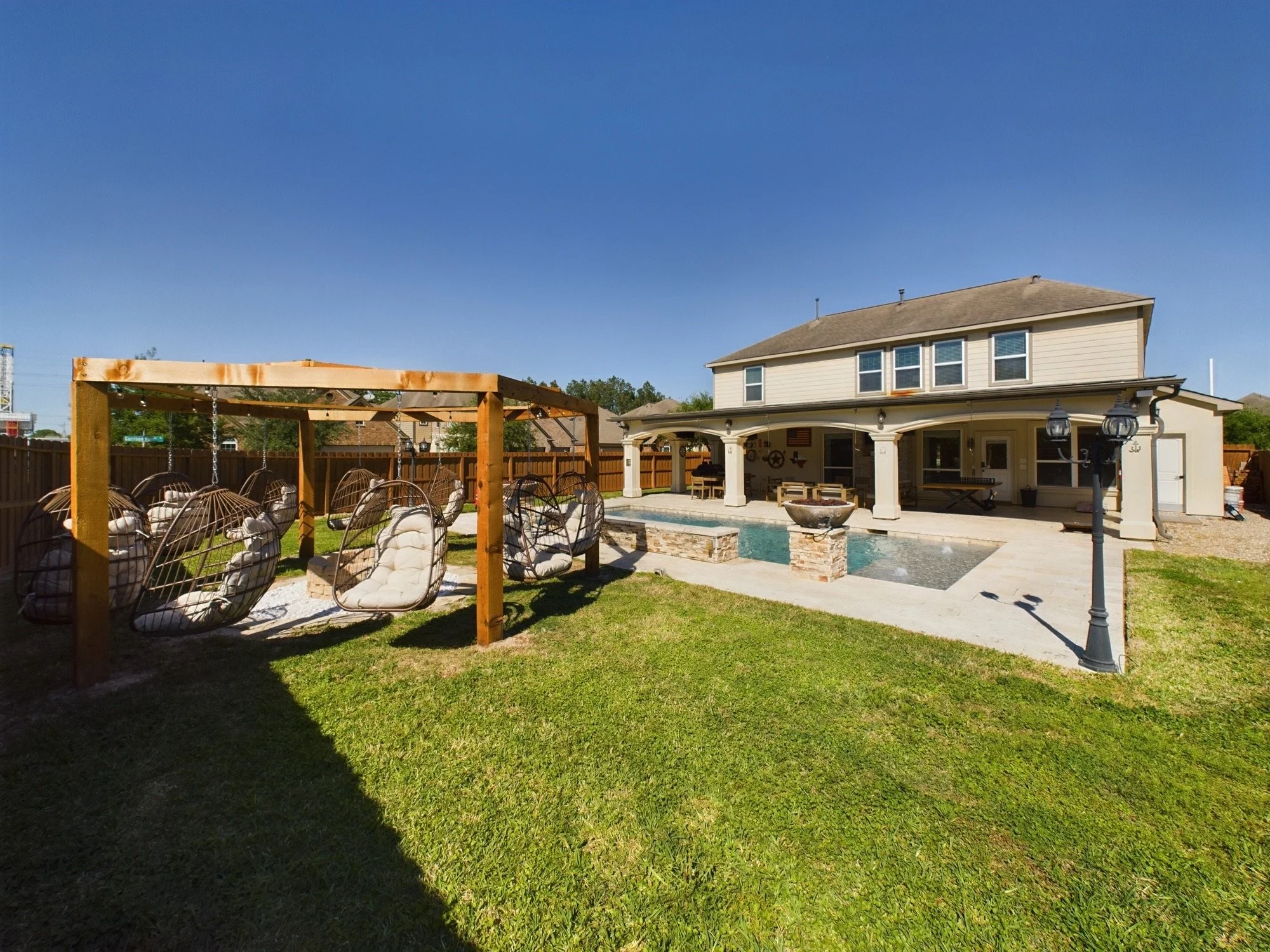 Backyard with a house, swimming pool, and grassy lawn. There is a wooden outdoor swing and a covered sitting area with outdoor furniture.