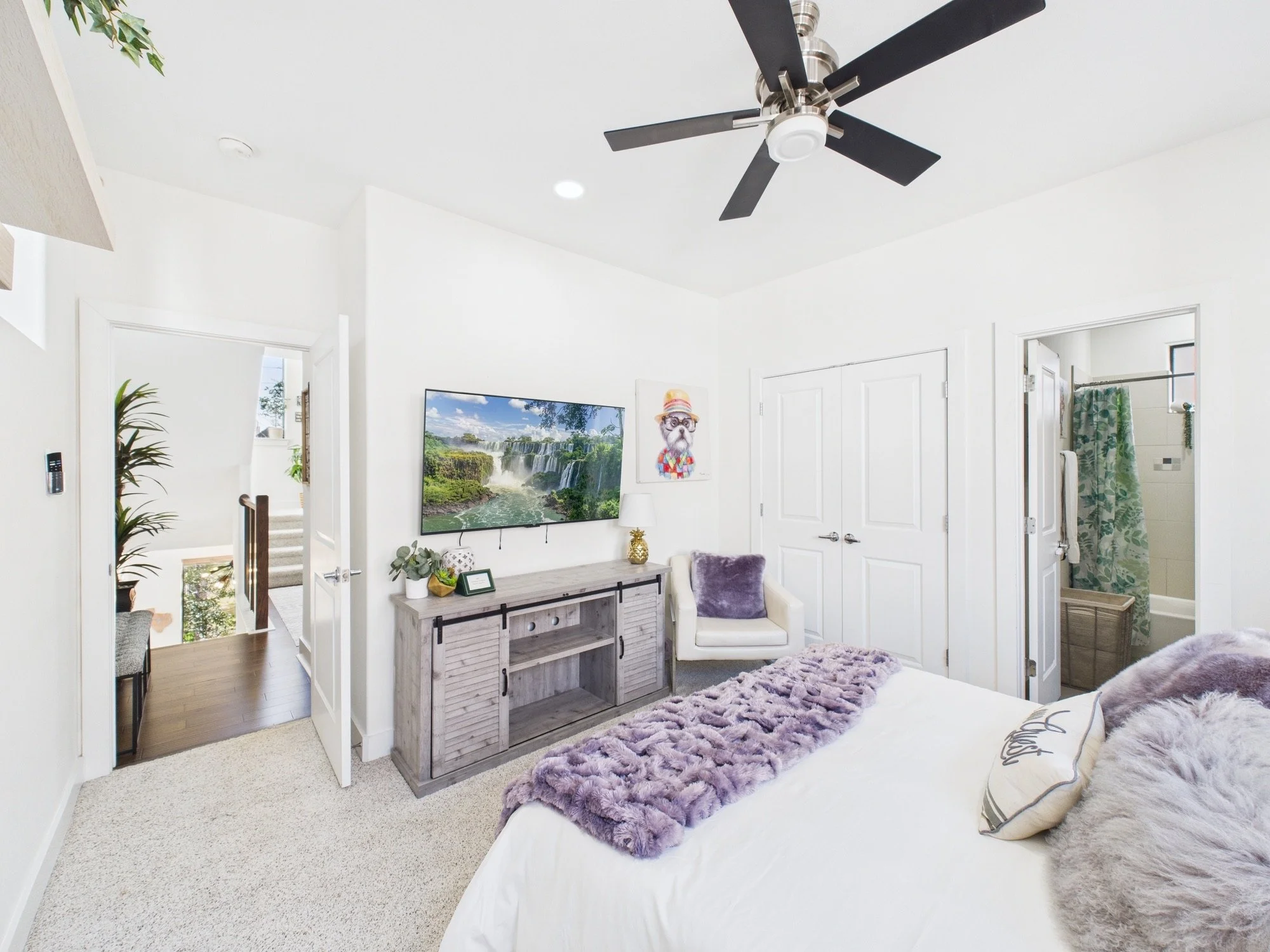 A bedroom with a bed, lavender plush blanket, and pillows, a white dresser beneath wall-mounted TV, framed print of a dog, ceiling fan, and a purple rug, with an en-suite bathroom visible through an open door.
