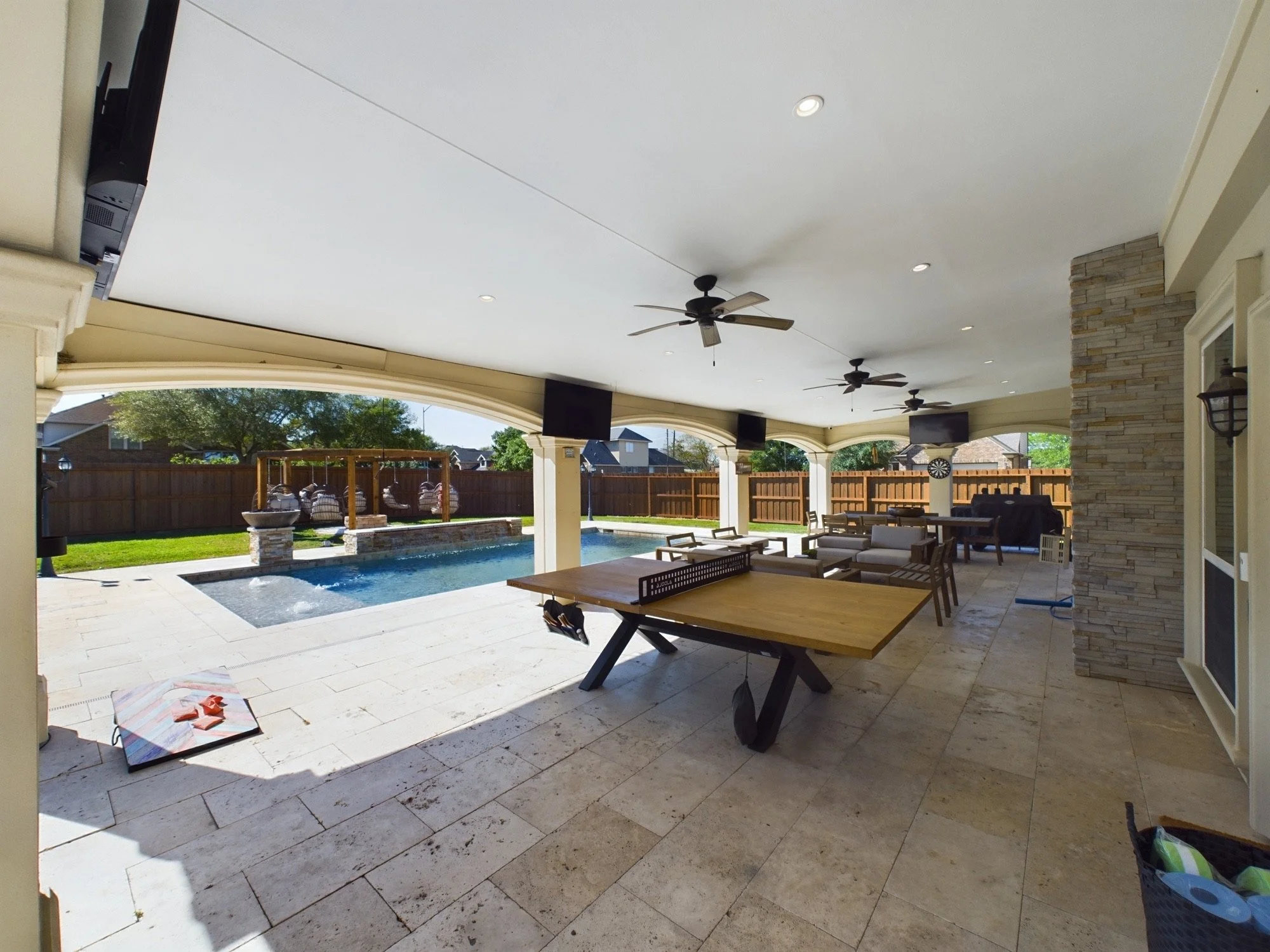 Covered backyard patio with ceiling fans, outdoor furniture, a swimming pool, and a fence, in a sunny backyard.