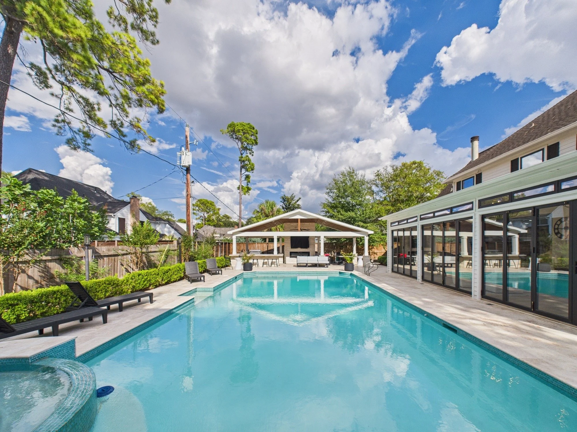 A backyard with a swimming pool, outdoor lounge chairs, a covered patio area, trees, and a house with large windows under a blue sky with clouds.