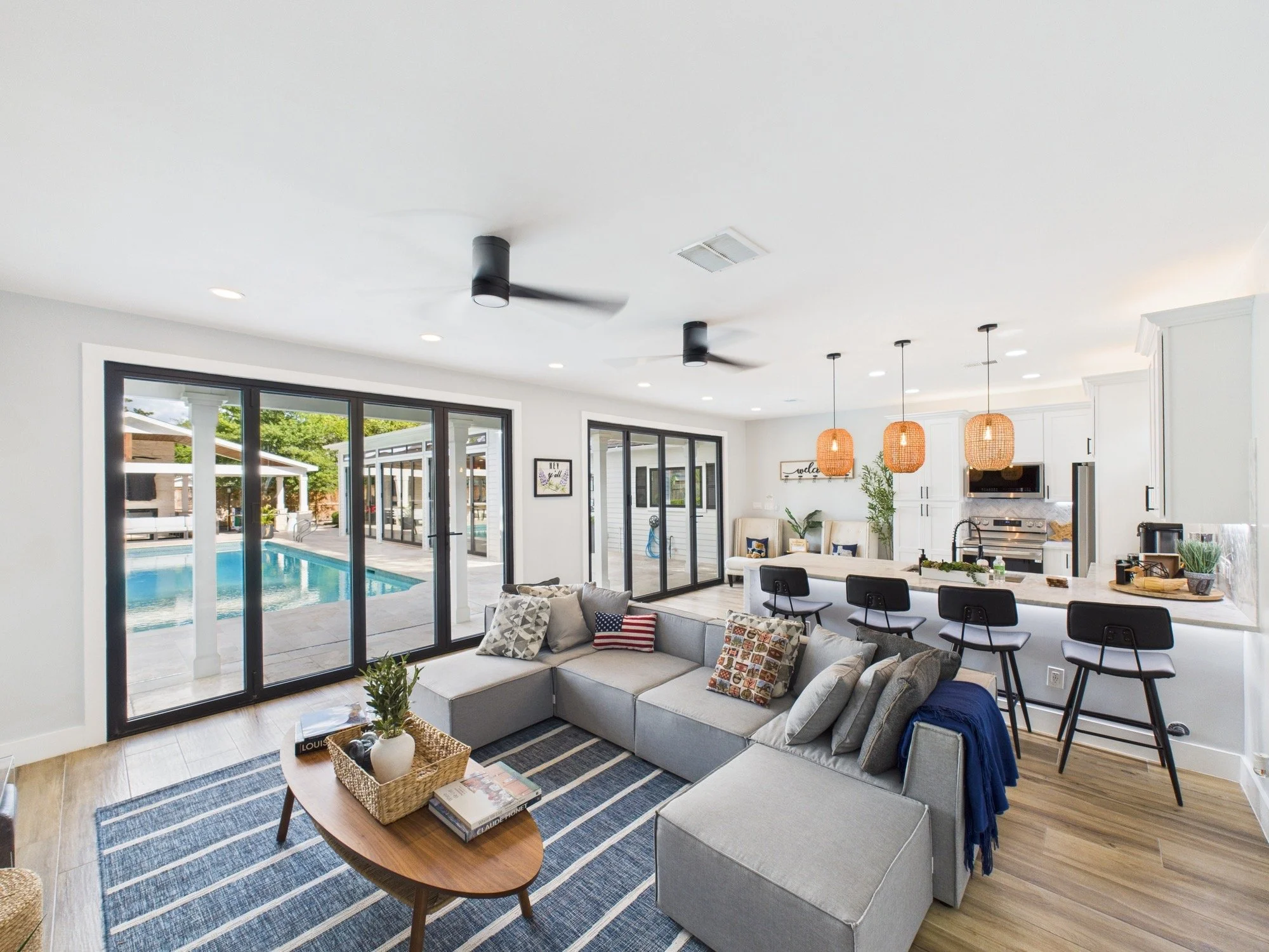 Open-concept living room with large sliding glass doors leading to a backyard pool, white walls, modern gray sectional sofa with patterned pillows, wooden coffee table with books, and a kitchen area with hanging orange pendant lights.