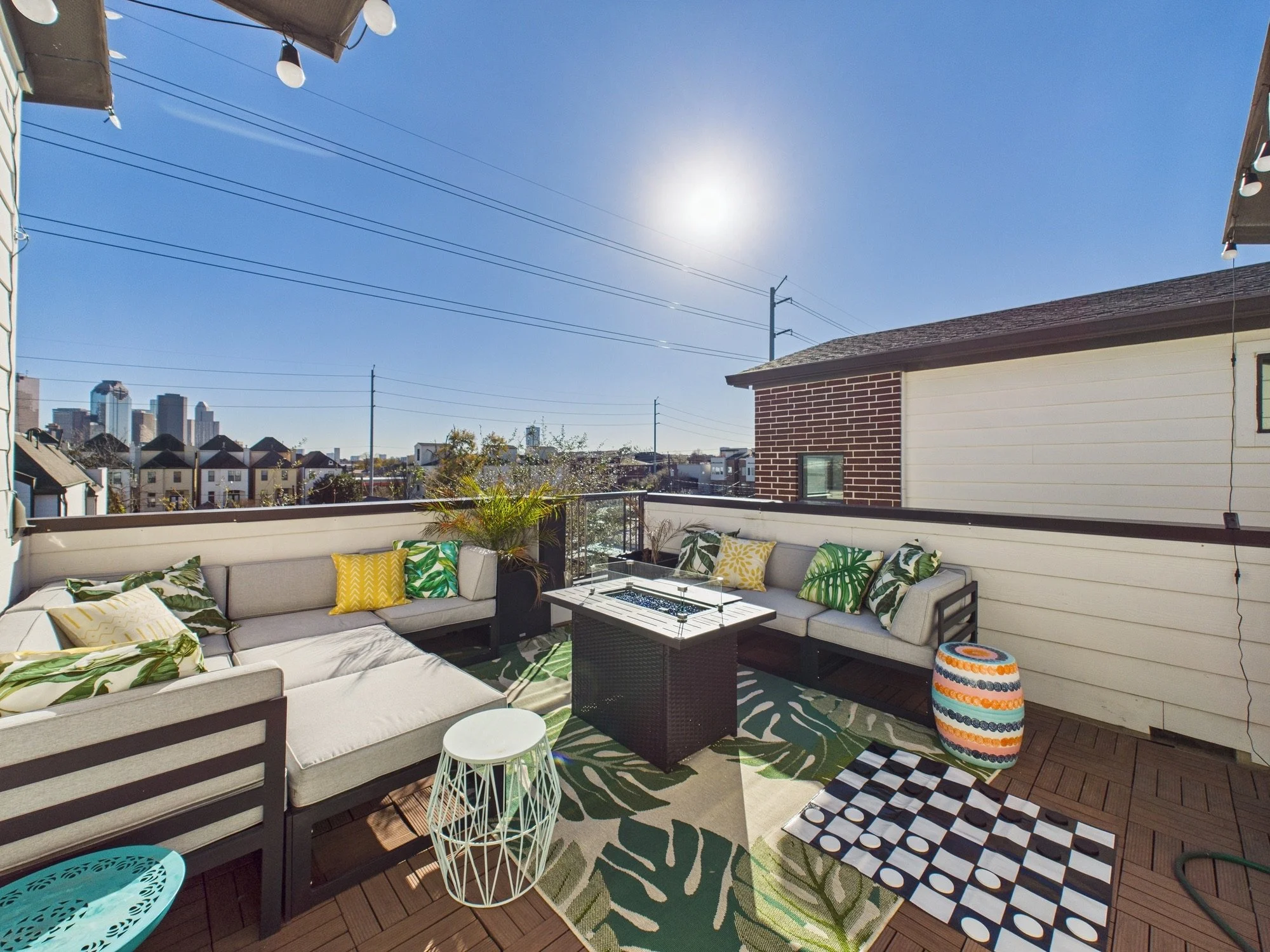 A rooftop patio with outdoor seating, colorful cushions, plants, and view of a city skyline with tall buildings under a clear blue sky with the sun shining.