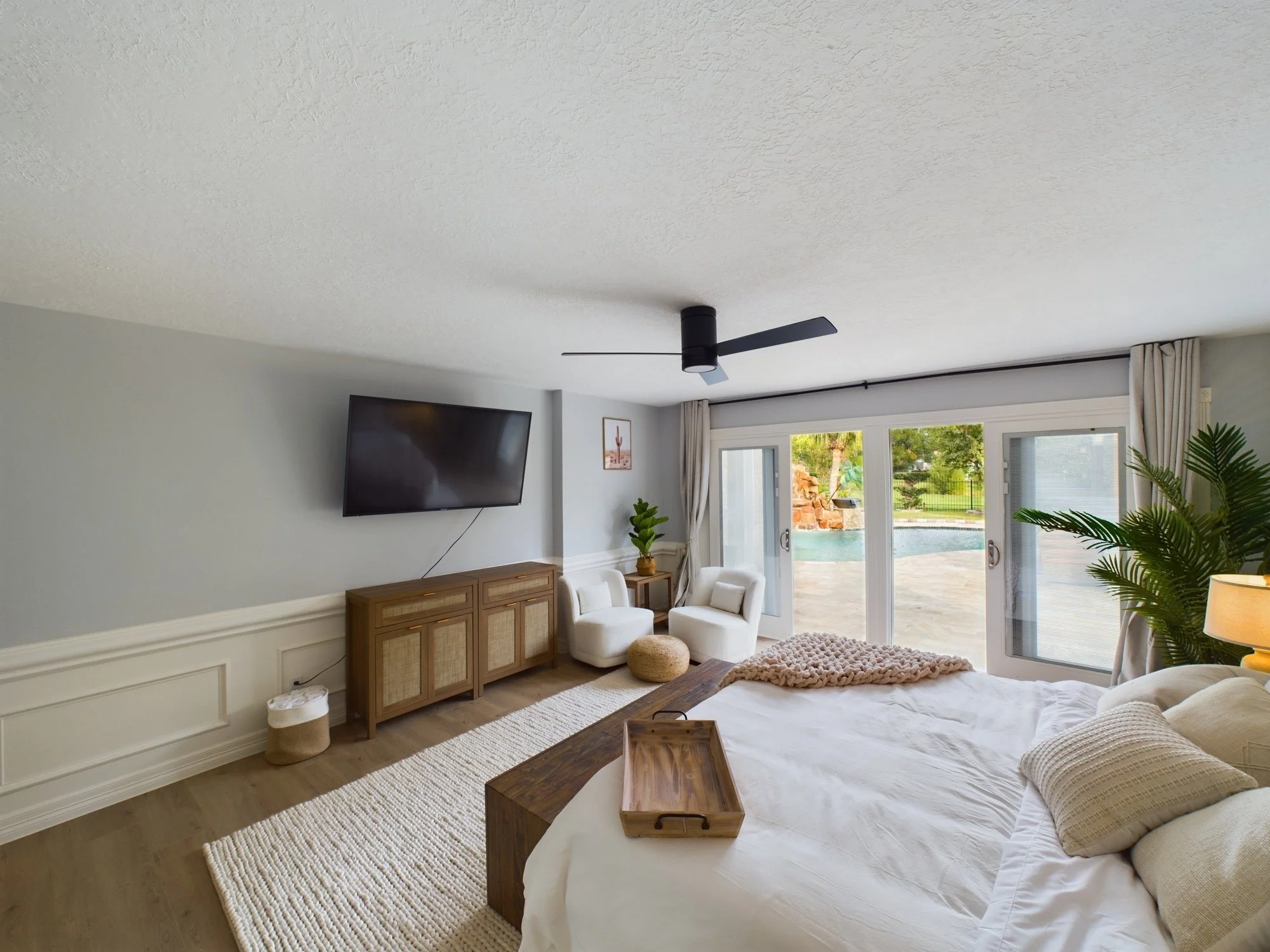 Modern living room with large glass doors opening to a backyard pool, beige curtains, a wall-mounted flat-screen TV, and a ceiling fan.