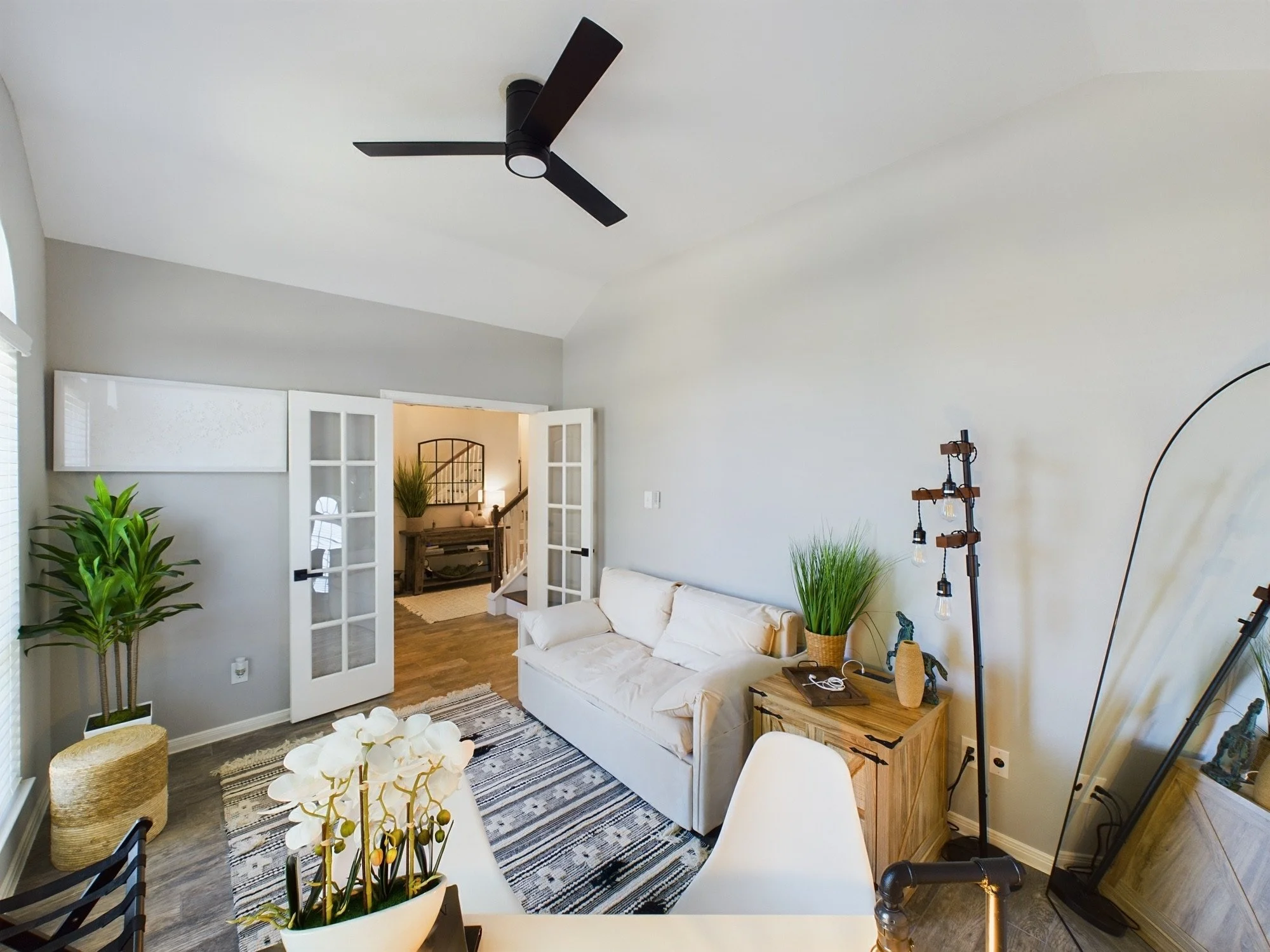 A living room with a white couch, wooden side table, potted plants, a black ceiling fan, and an open doorway leading to a hallway with a mirror and stairs.