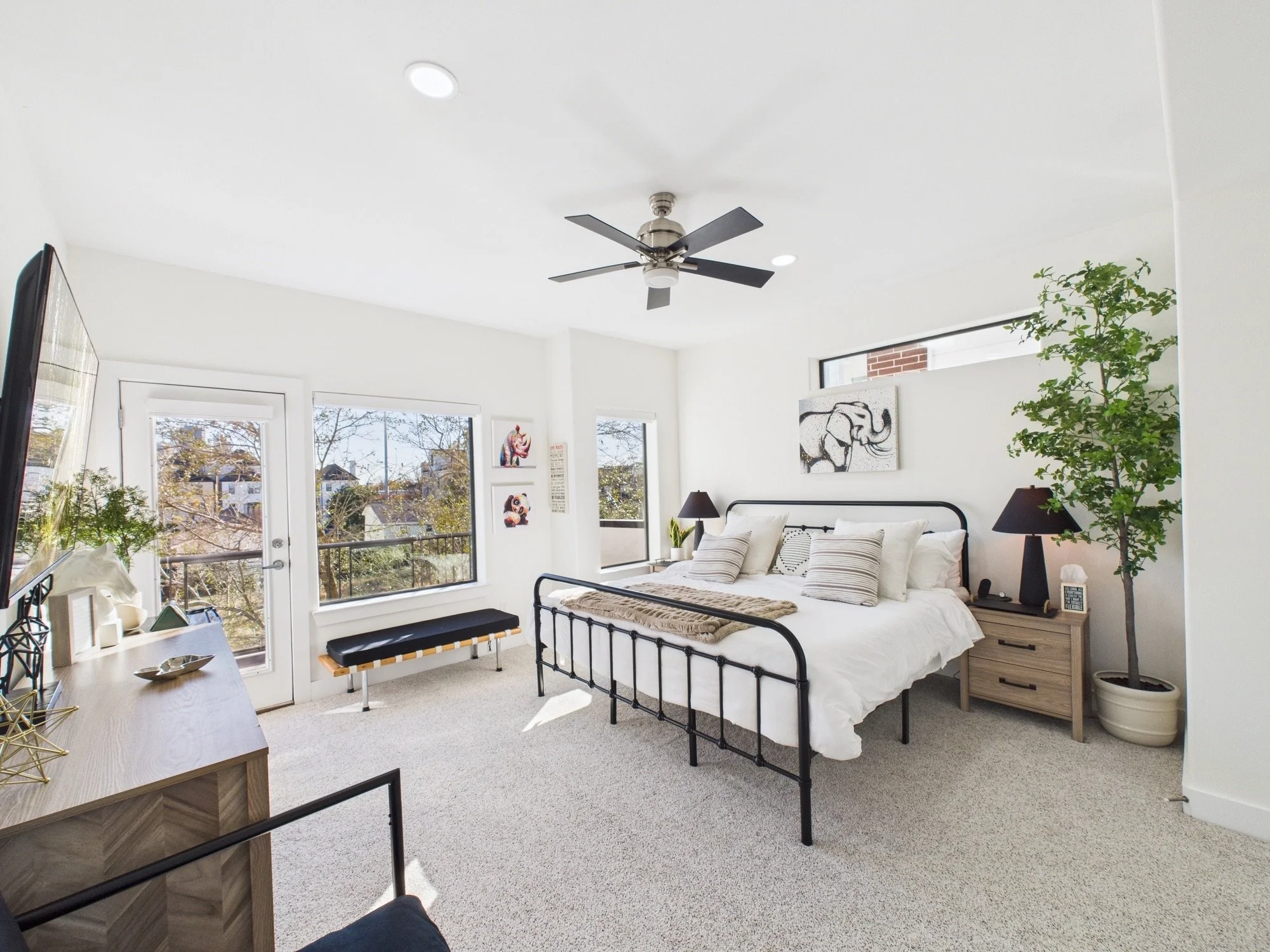 Bright bedroom with white walls, large windows, a black metal bed frame with white bedding, two black bedside lamps, a wooden nightstand, a tall green plant, and wall art including an elephant and a panda, with a ceiling fan.