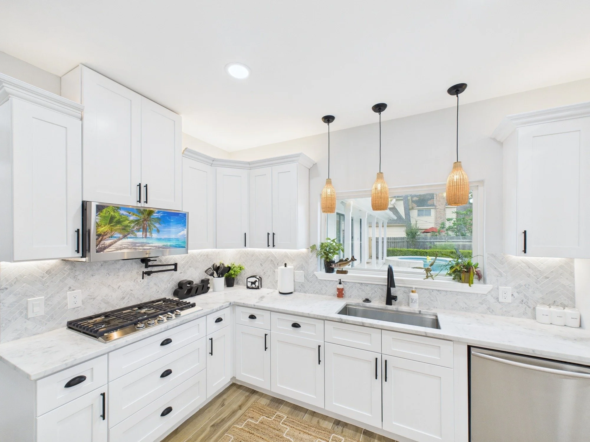 A modern kitchen with white cabinets, a marble countertop, a gas stove, a sink, hanging wicker pendant lights, and a window overlooking a backyard with a pool and trees.