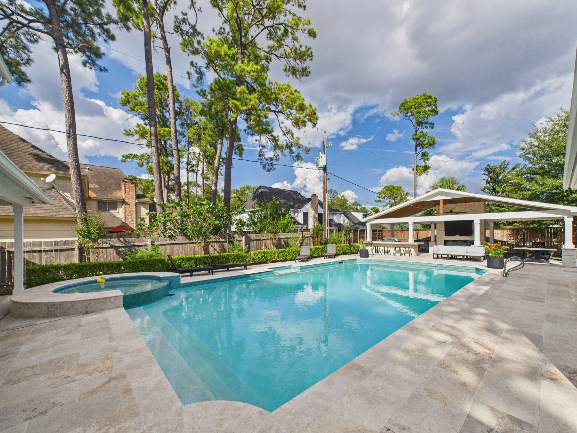 Residential backyard swimming pool with a hot tub, poolside lounge chairs, and a shaded outdoor seating area, surrounded by a wooden fence and tall trees with cloudy sky overhead.