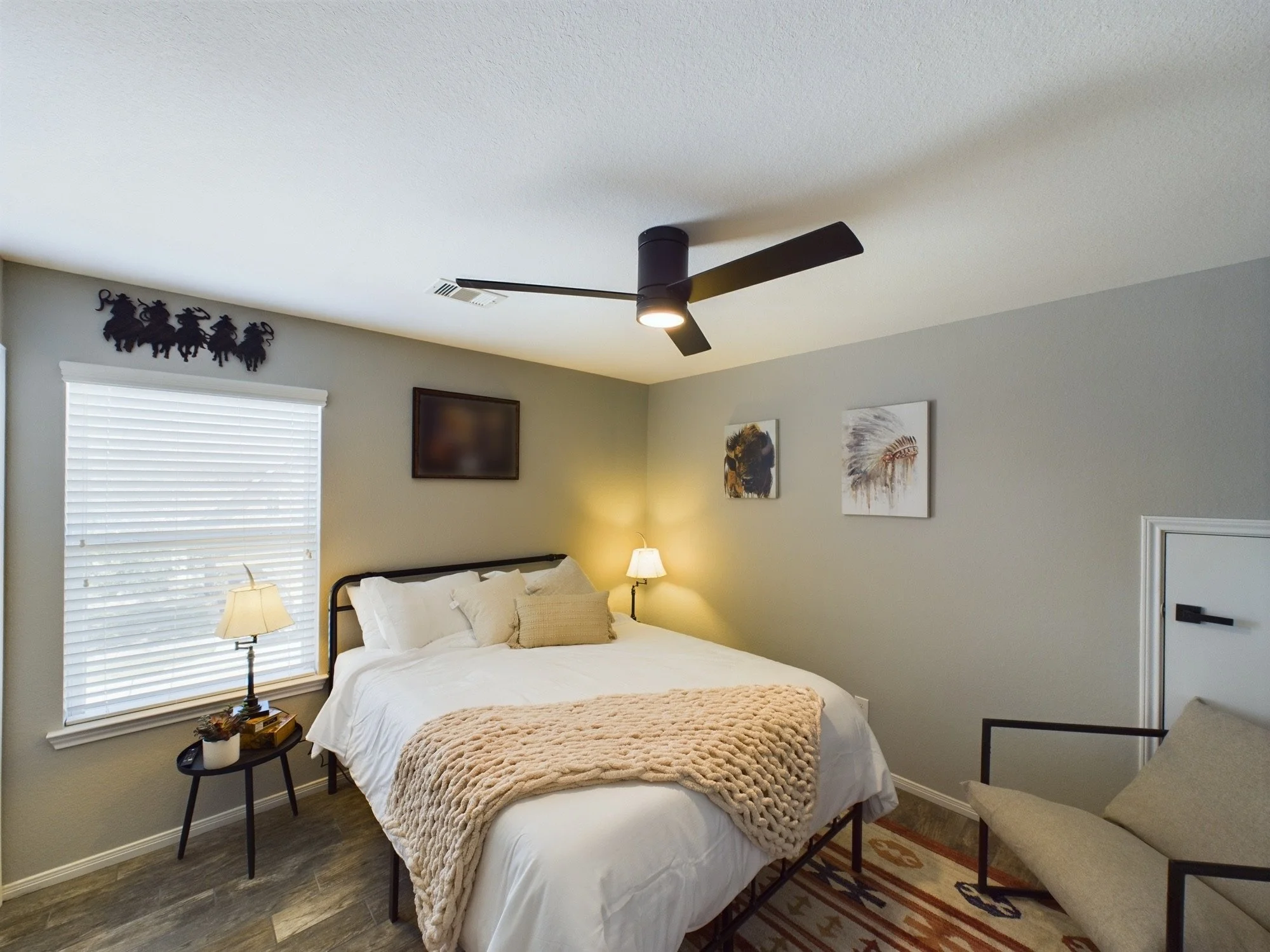 A bedroom with two twin beds pushed together, each with white bedding and beige knitted throws, a small black bedside table with a lamp and decorative items, a window with white blinds, wall art including animal prints and a wall decoration of seven 