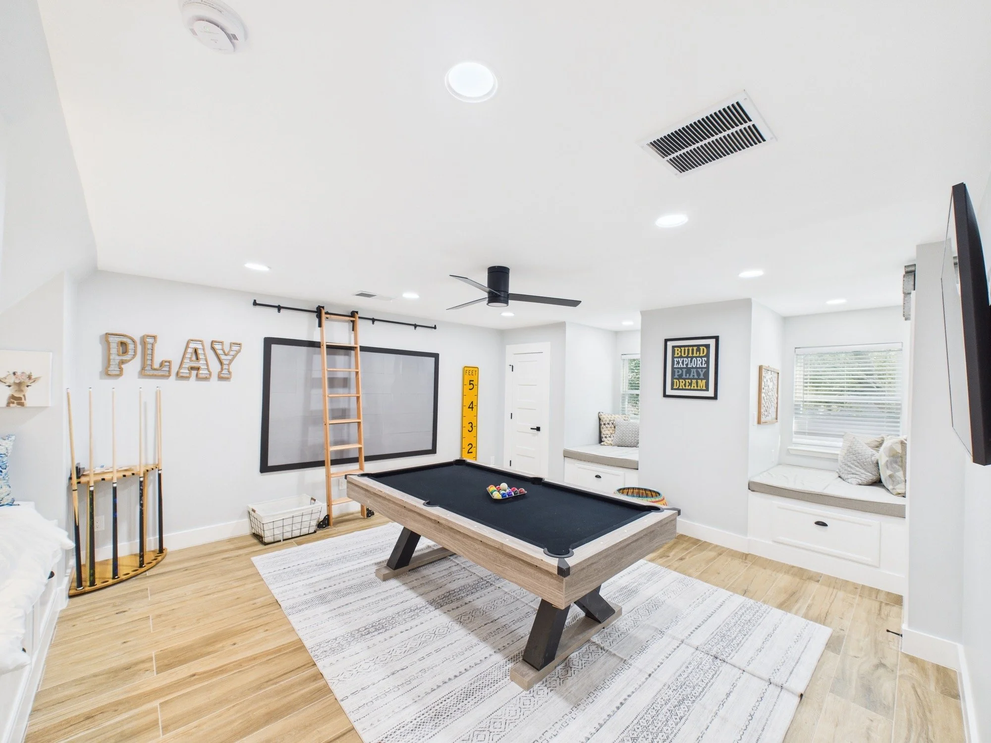 A recreation room with a black pool table, light wood flooring, a white rug, and a white wall decorated with framed posters, including one reading "Build Explore Play Dream." There is a snack basket and a golf club rack nearby, and windows with blind