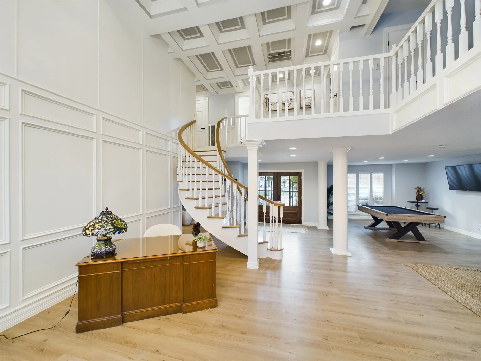 Bright living room with white walls and wooden flooring, featuring a staircase with a wooden handrail, a wooden console table with a stained glass lamp, and a pool table in the background.