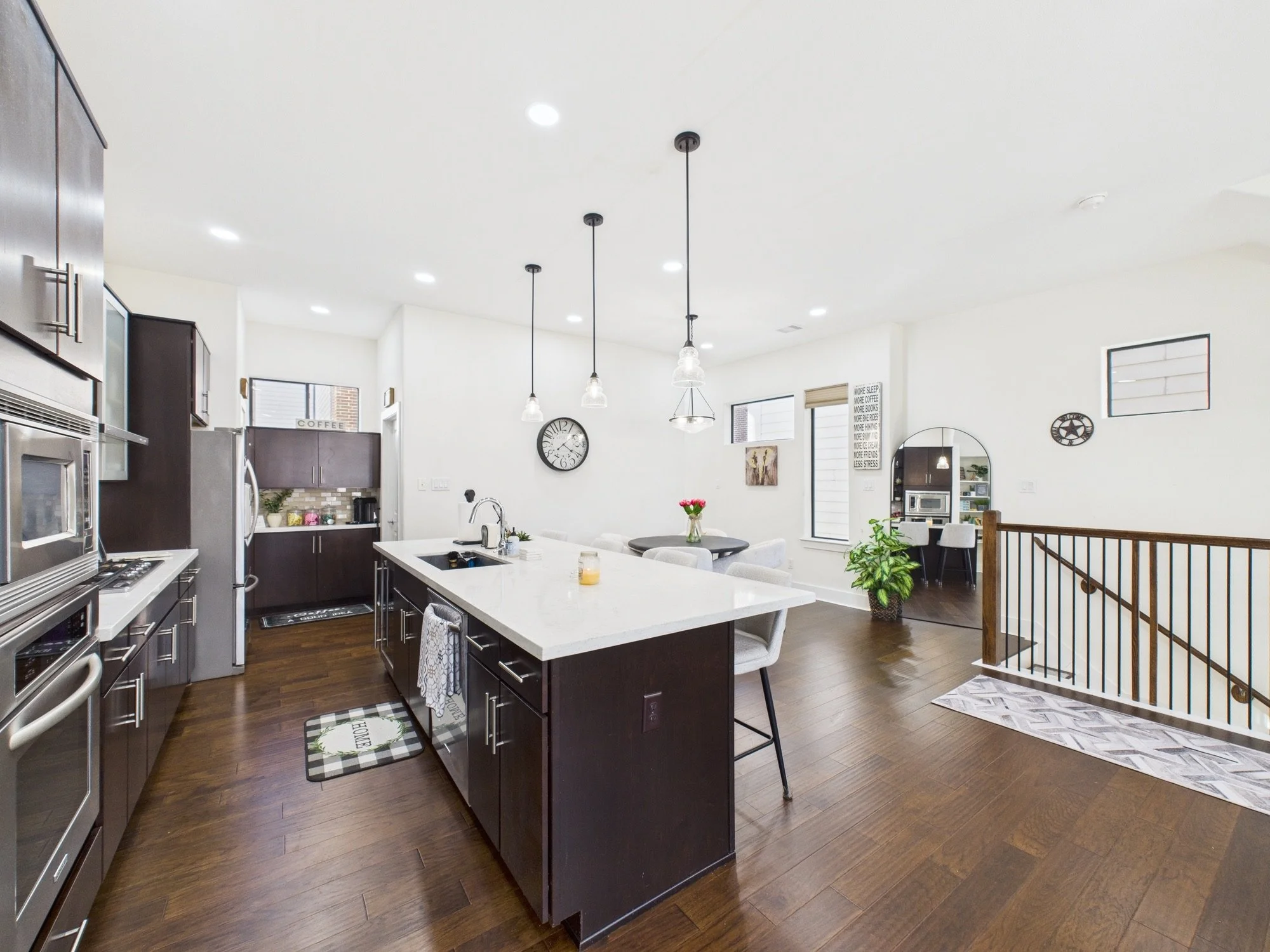 A modern kitchen and dining area with a large white island, dark wood cabinets, stainless steel appliances, hardwood floors, and pendant lighting fixtures hanging from a white ceiling.