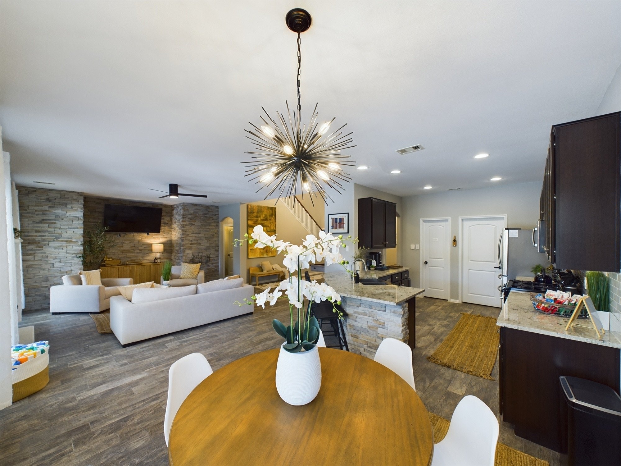 A modern living room with a kitchen in the background, featuring white sofas, a wooden dining table with white chairs, a large decorative chandelier, and a vase with white flowers on the table.