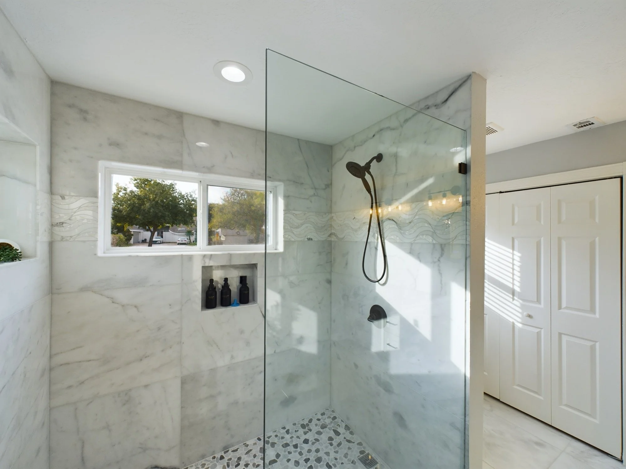 Modern walk-in shower with marble walls and a glass panel. There is a window letting in natural light, and a showerhead with a handheld attachment. Storage bottles are on a built-in shelf.