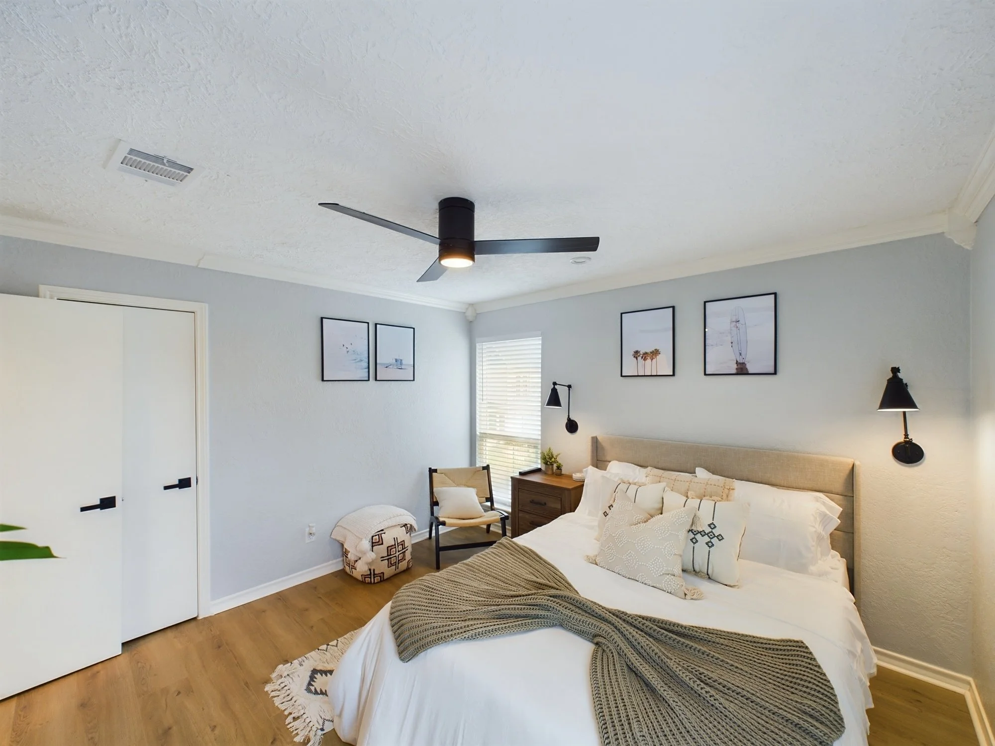 A bedroom with a bed, a wooden nightstand, framed pictures on the wall, and a ceiling fan with lights.