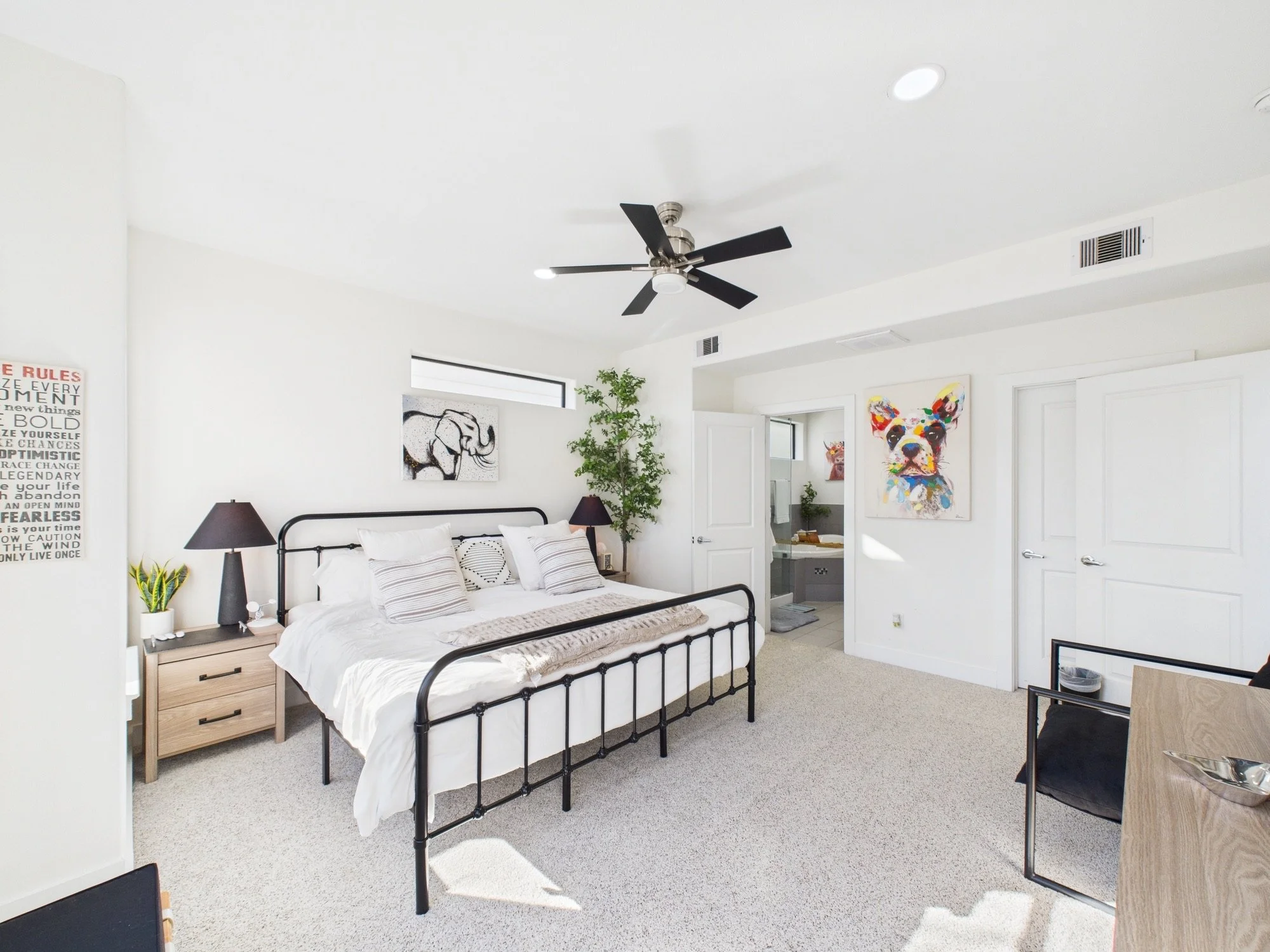 Bright bedroom with white walls, black bed frame, white bedding, two black lamps, artwork of bulldog and elephant, indoor potted plant, and doorway leading to a bathroom.