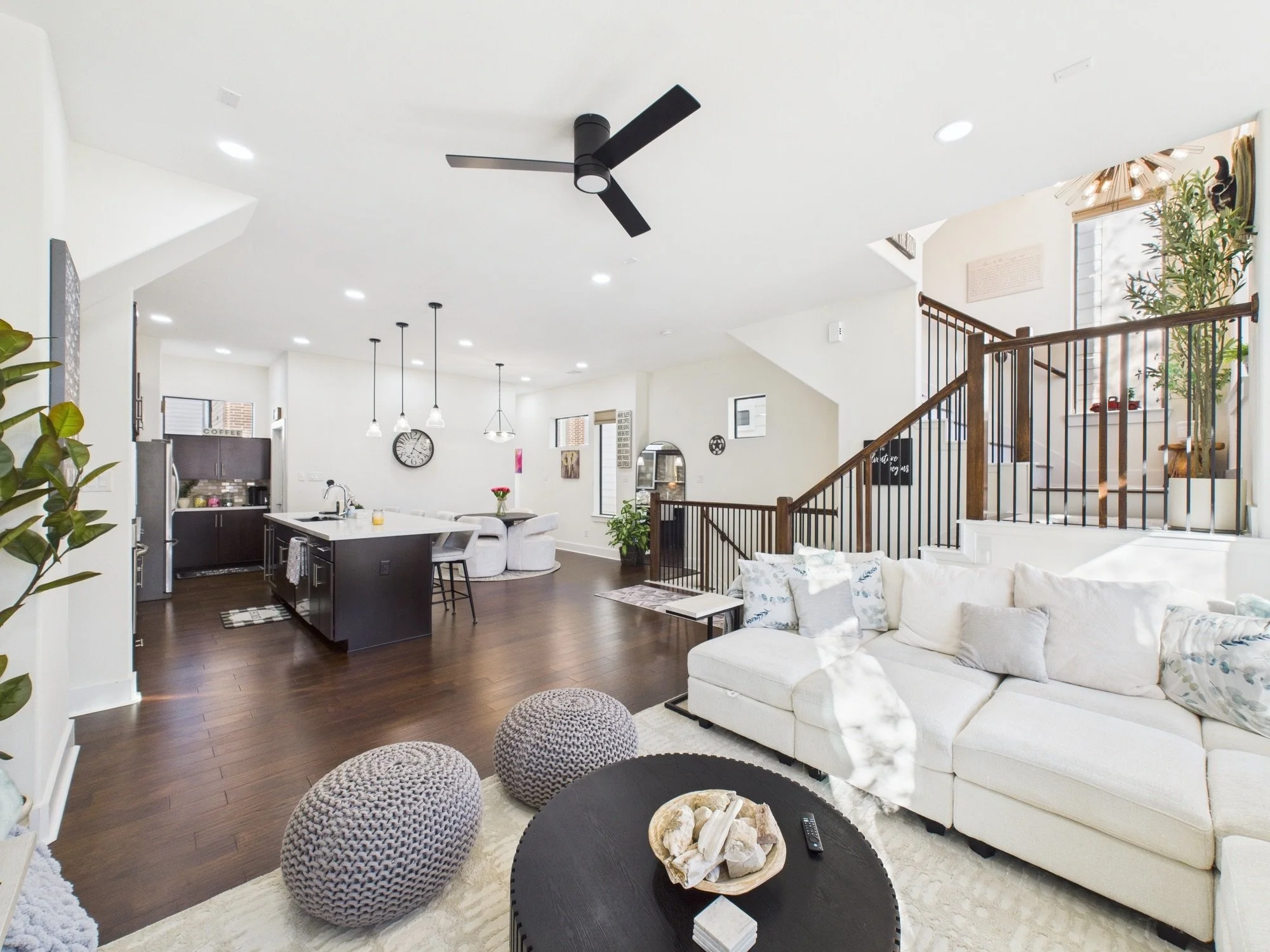 Open-concept living room and kitchen with modern decor, white walls, dark hardwood floors, black ceiling fan, staircase with wooden railing, and large windows letting in natural light.