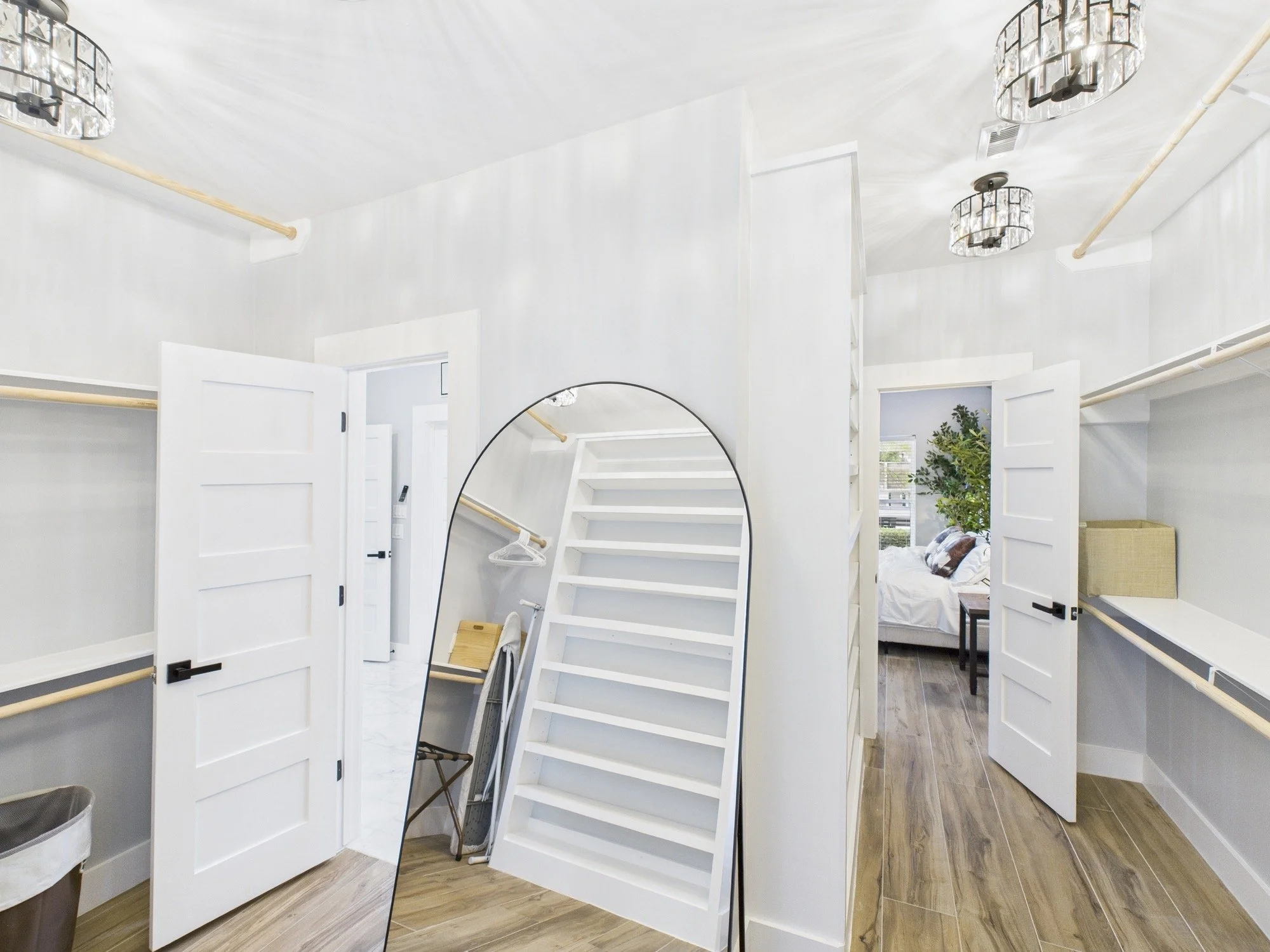 A room with white walls, ceiling, and doors, featuring two chandeliers, wooden rods on the walls, a mirror on a stand, a small chair, and a bed with pillows and a plant nearby.