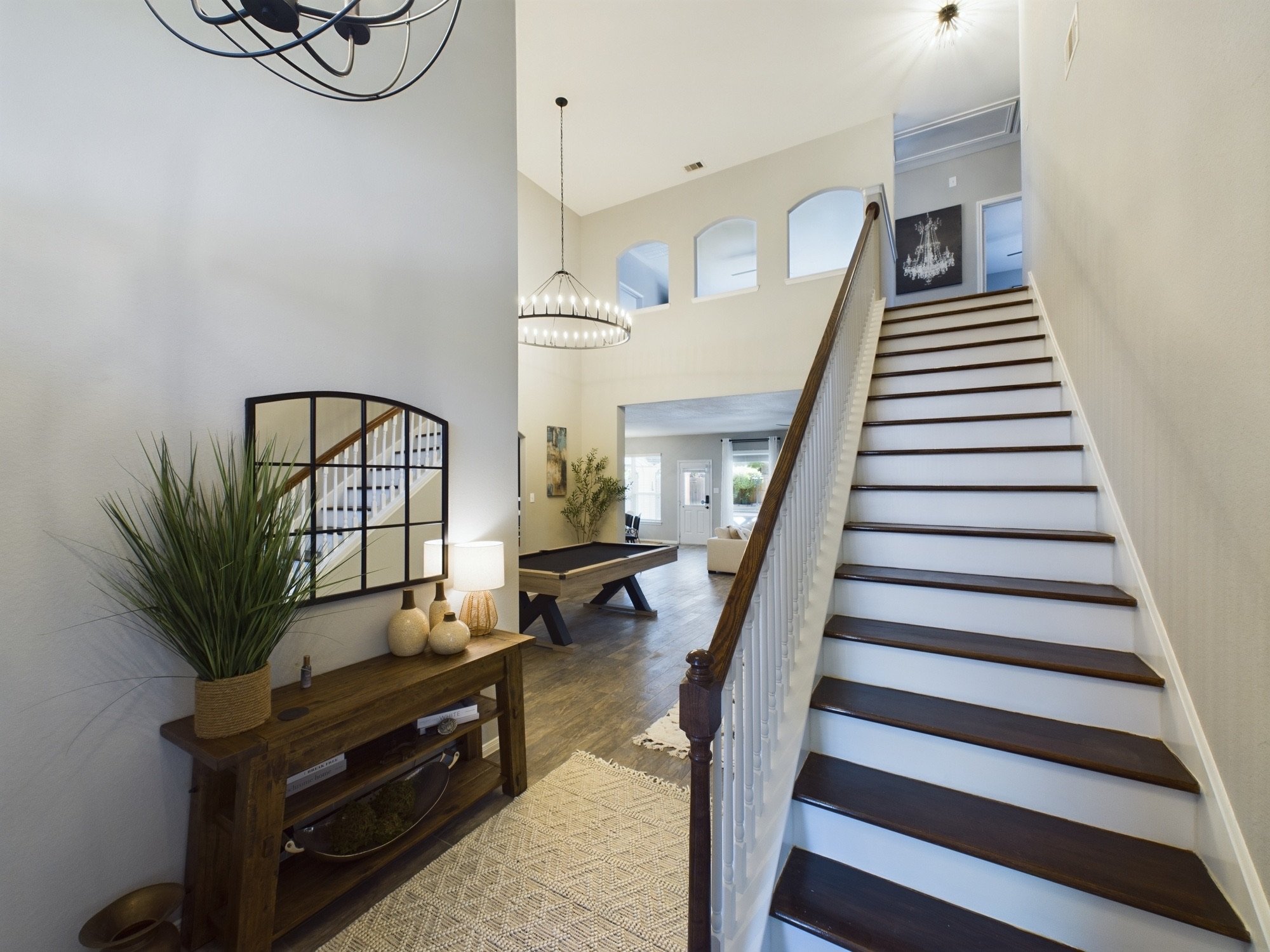 Interior of a modern house entryway showcasing a staircase with dark wooden steps and white risers, a hallway with a pool table, a wall with a chandelier painting, and a decorative console table with vases and a mirror, illuminated by natural light f