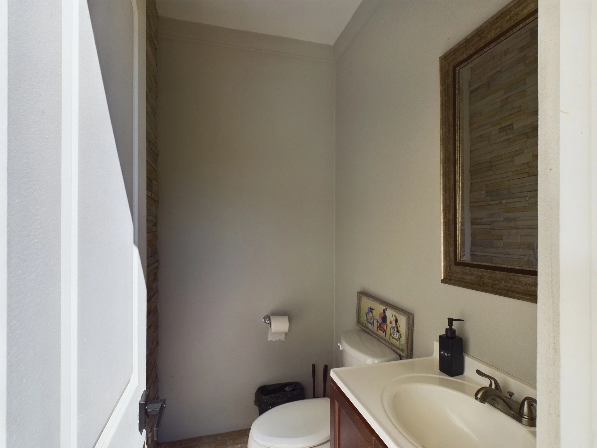 Small bathroom with a white sink and a dark wooden cabinet beneath it, a mirror above the sink, a framed picture on the wall, a toilet with a box of soap on top, a toilet paper roll on a holder, and a small trash can.