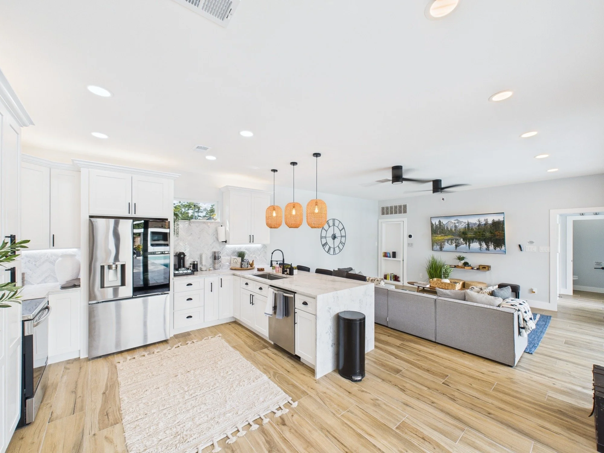 Modern open-concept kitchen and living room with white cabinetry, marble island, hanging pendant lights, and wall-mounted TV, with a ceiling fan and wood flooring.