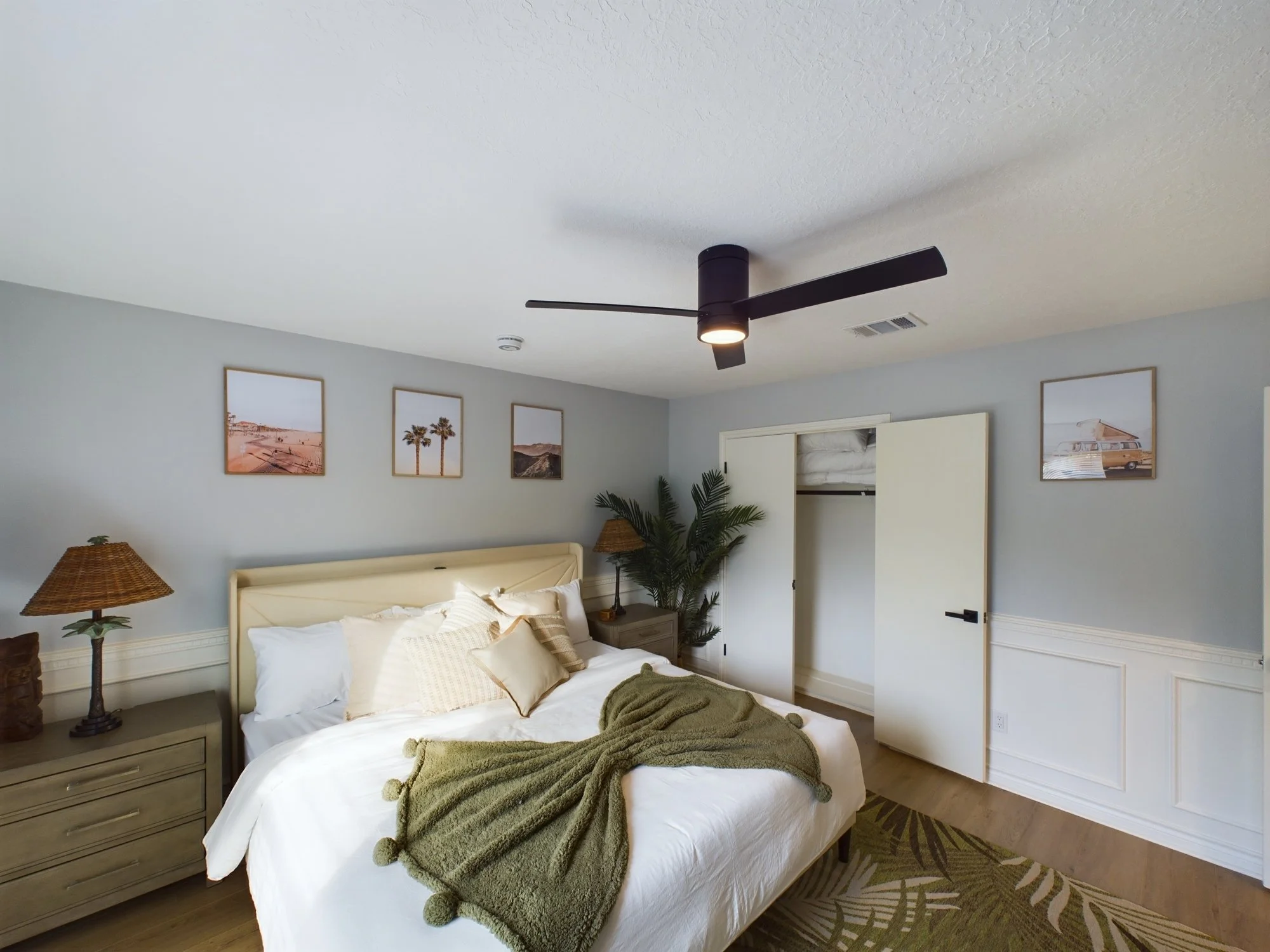 A bedroom with a white bed, green throw blanket, bedside table with lamps, framed landscape photographs on the wall, a closet with sliding doors, ceiling fan, and a patterned rug.