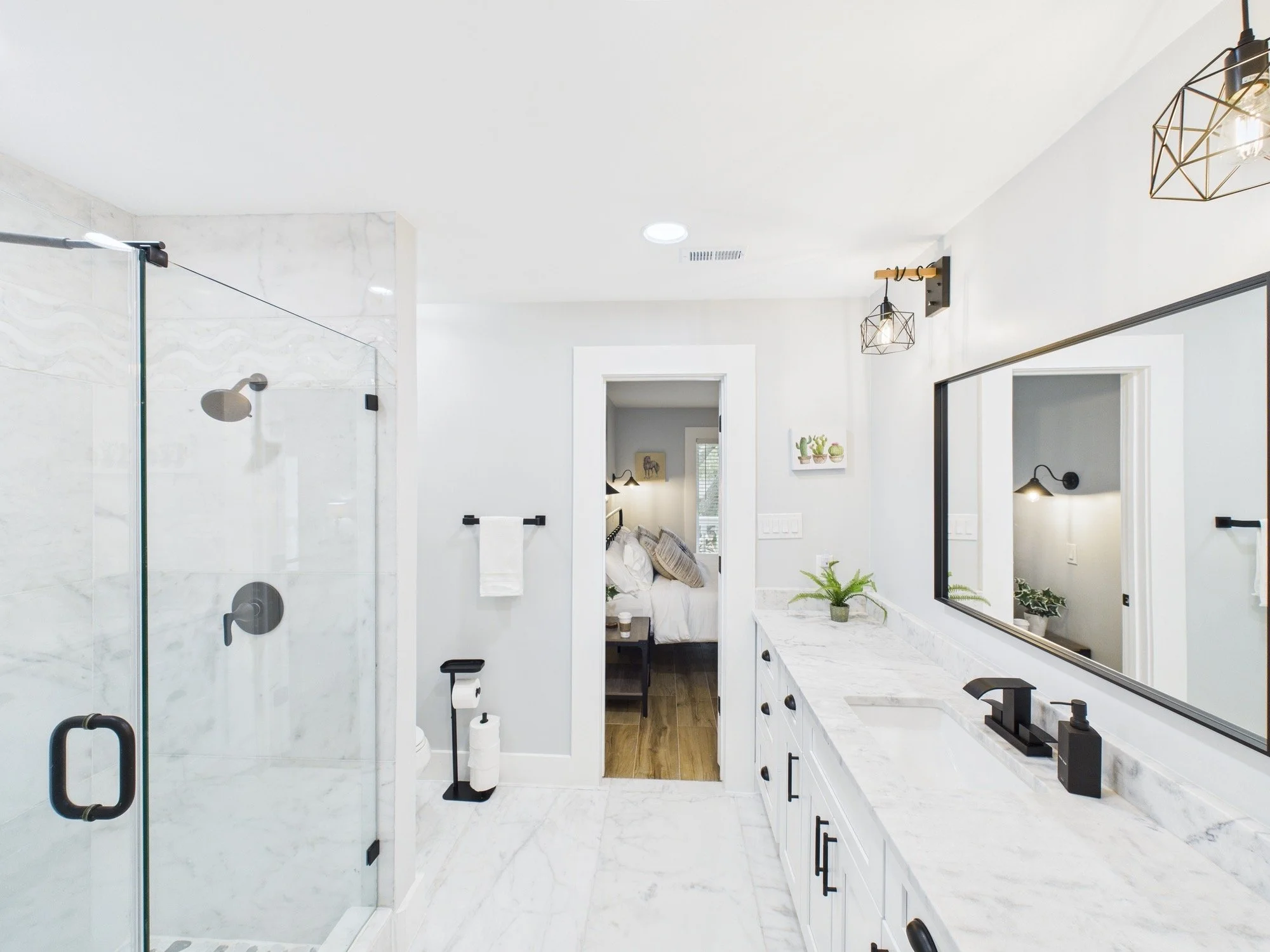 Modern bathroom with a walk-in shower with glass door, white marble walls, black fixtures, and a white vanity with a marble countertop, mirror, and decorative plants.