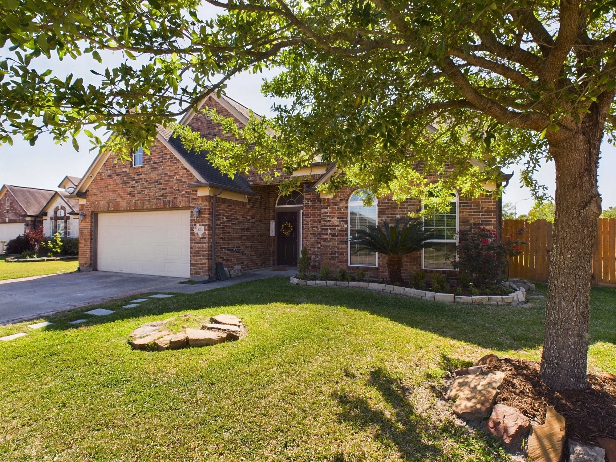 A brick house with a garage, front door, and windows, partially obscured by a large leafy tree in the front yard, with lush green grass and a decorative plant near the house, under a clear blue sky.