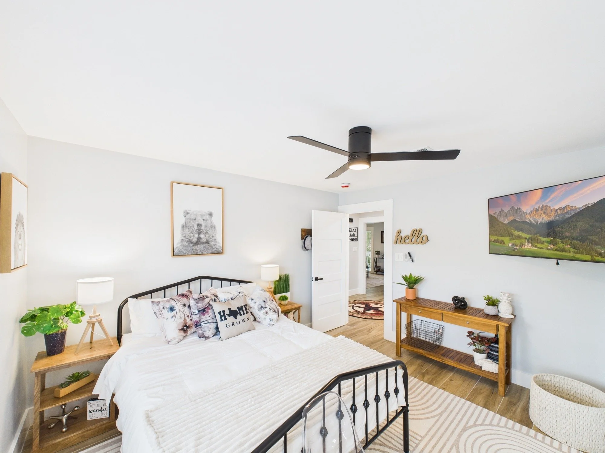 Modern bedroom with a black metal bed frame, white bedding, and decorative pillows. Wooden nightstand with a potted plant, framed dog artwork on the wall, and large landscape picture. Ceiling fan and a door leading to another room.