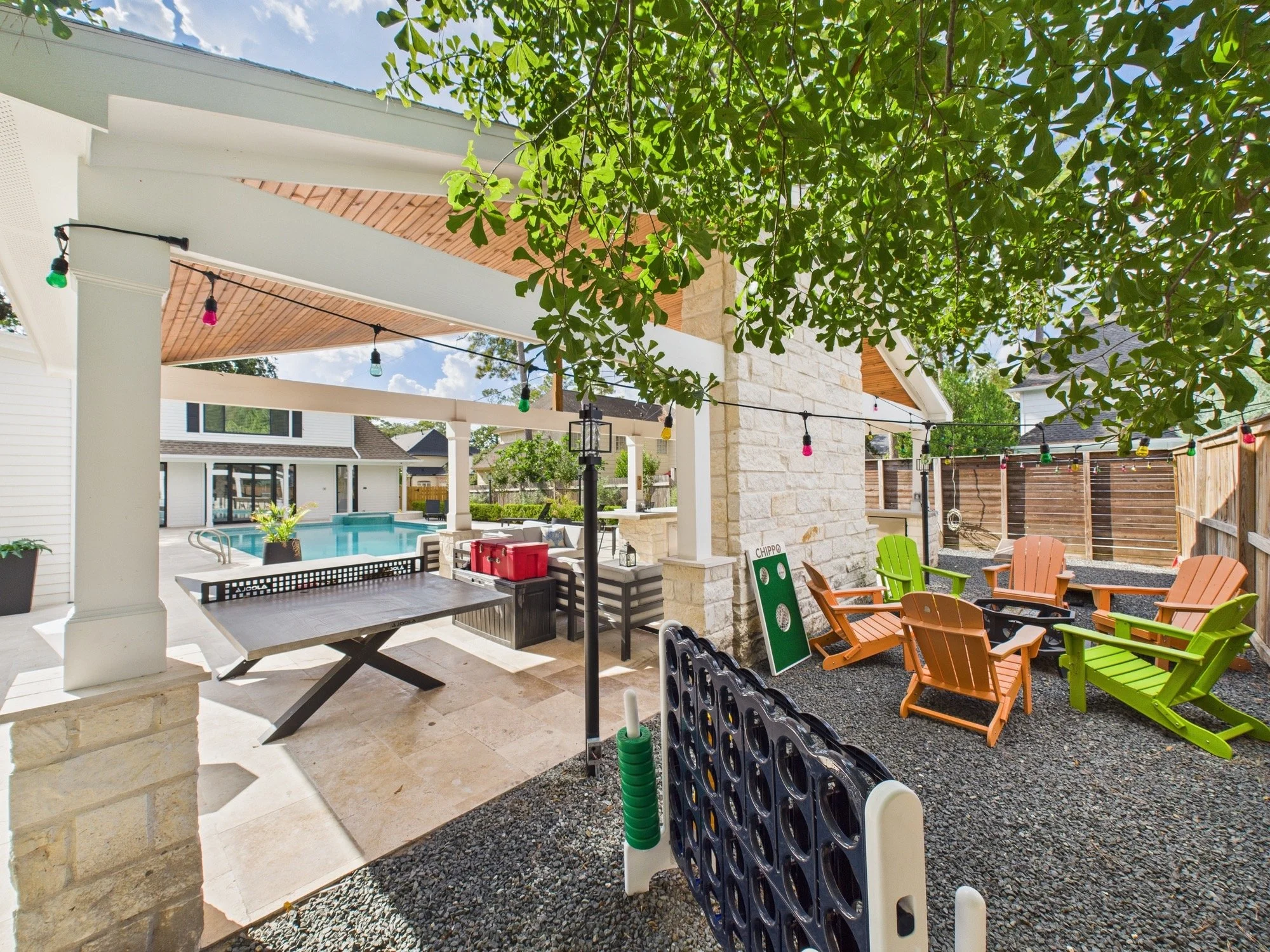 A backyard patio area with a covered seating area, string lights, a pool in the background, colorful Adirondack chairs, a small green cornhole game, and lush green trees overhead.