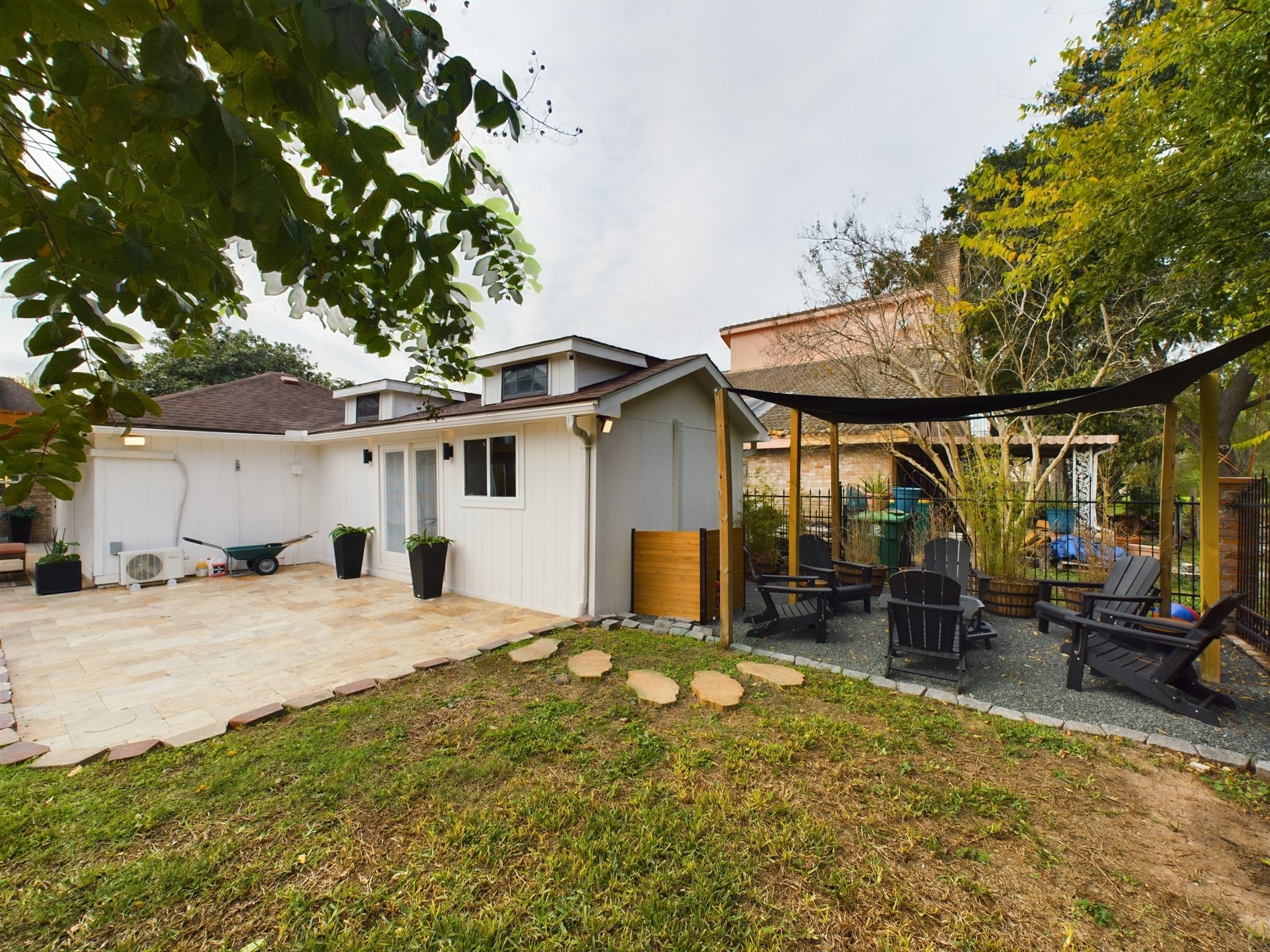 A backyard patio area with a light-colored stone floor, a white shed, outdoor furniture, potted plants, a shade sail, and trees with leaves in early fall.