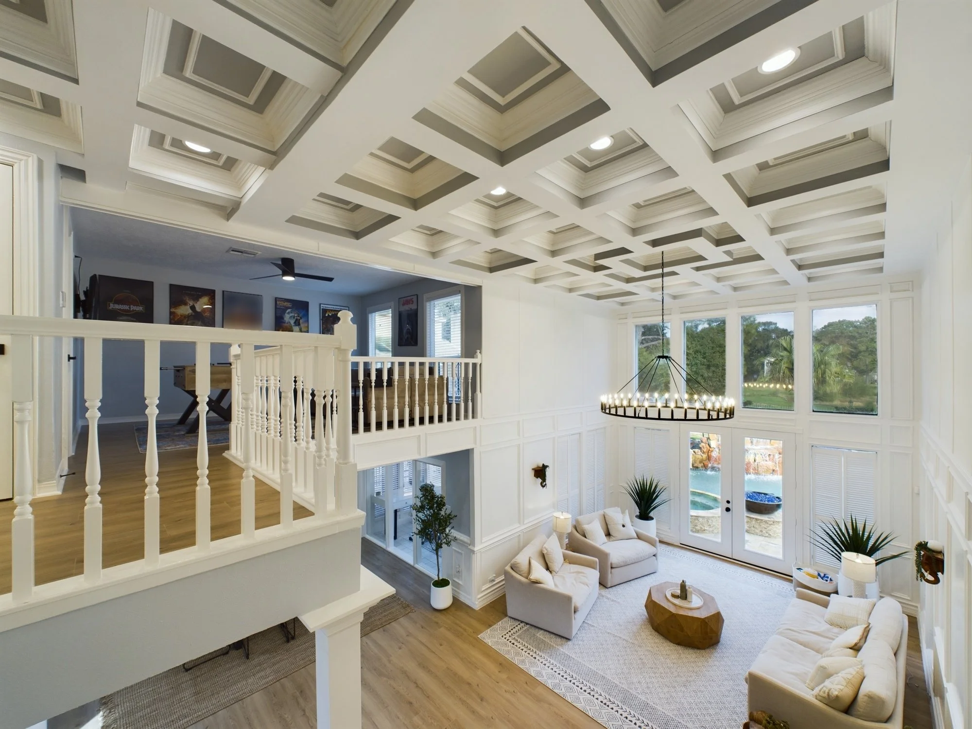 Bright living room with high vaulted ceiling featuring recessed lighting and a modern chandelier. Large windows and sliding glass doors provide natural light and views of greenery outside. The interior has white walls, beige furniture, and wooden flo