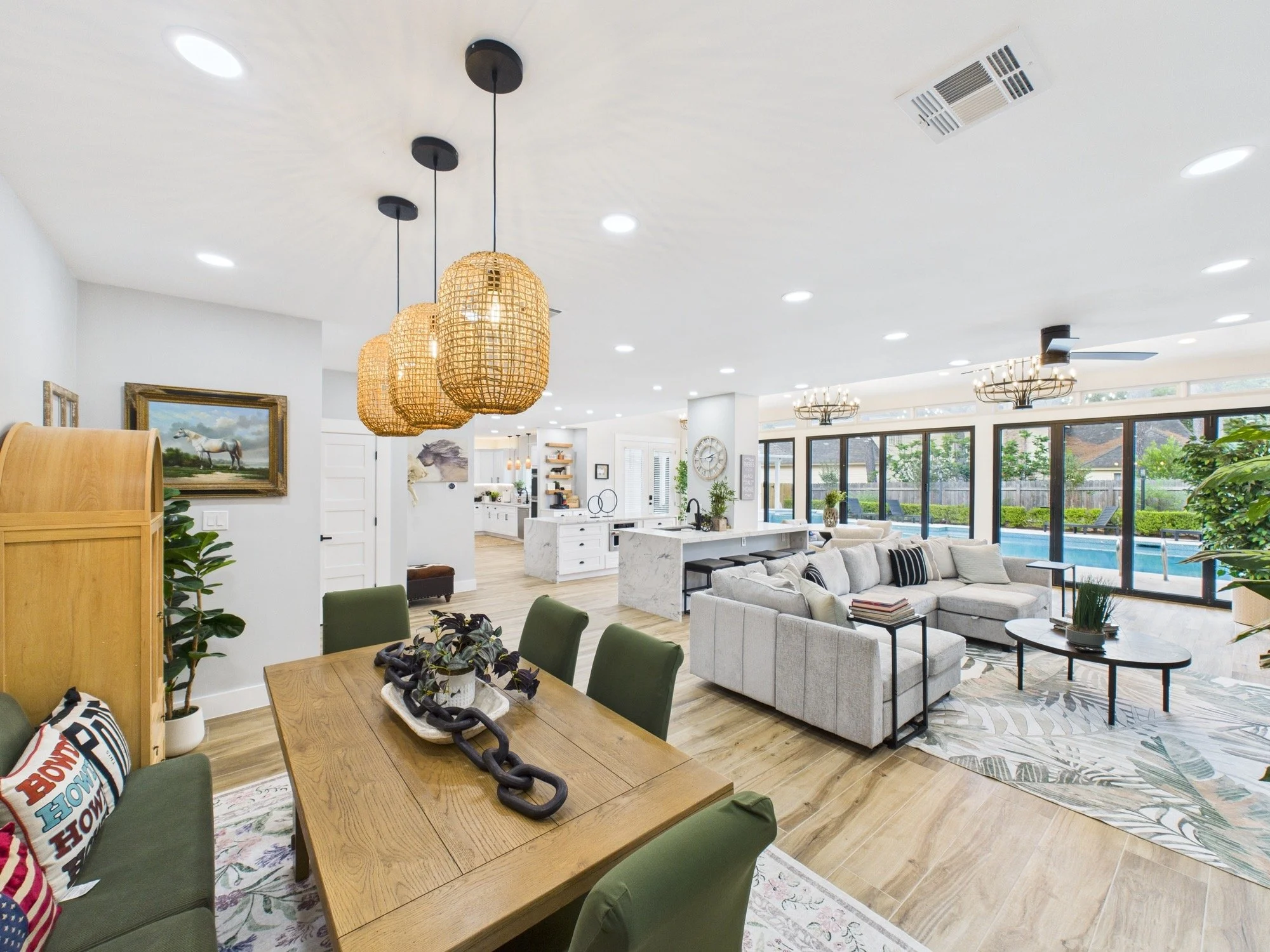 Open-concept living and dining area with white walls, wooden flooring, modern hanging light fixtures, sectional sofa, and large windows revealing a backyard with trees and outdoor furniture.
