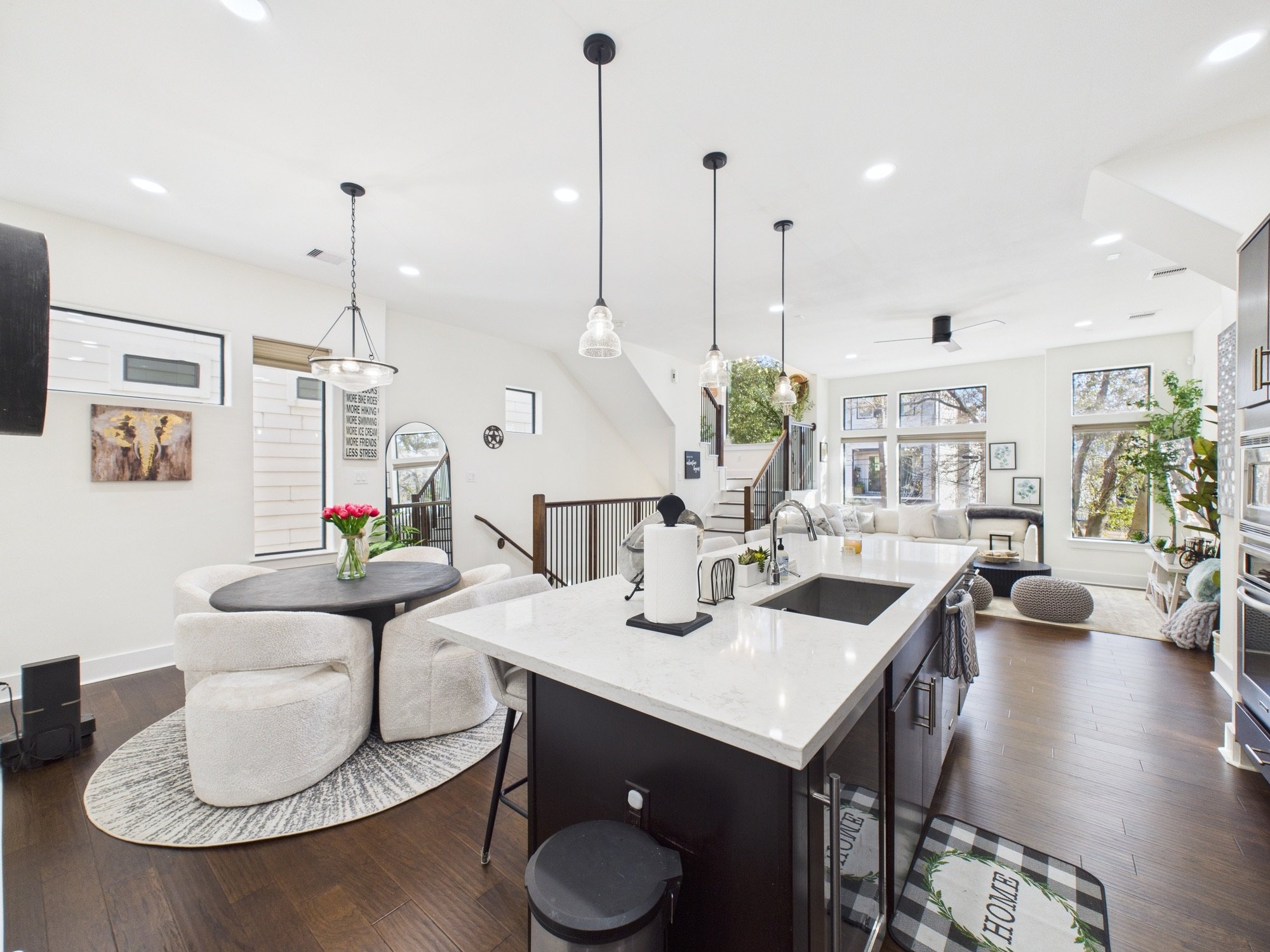 Open concept living space with white walls, large windows, dark hardwood floors, a kitchen island with a white countertop, and modern pendant lights hanging from the ceiling. There is a dining area with a round table and chairs, and a cozy sitting ar