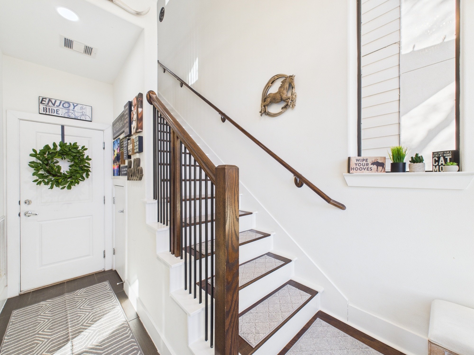 Interior view of a staircase with a wooden handrail and white steps, next to a white wall decorated with a horse wreath and framed artwork, a window ledge with plants and signs, and a closed white door with a green wreath.