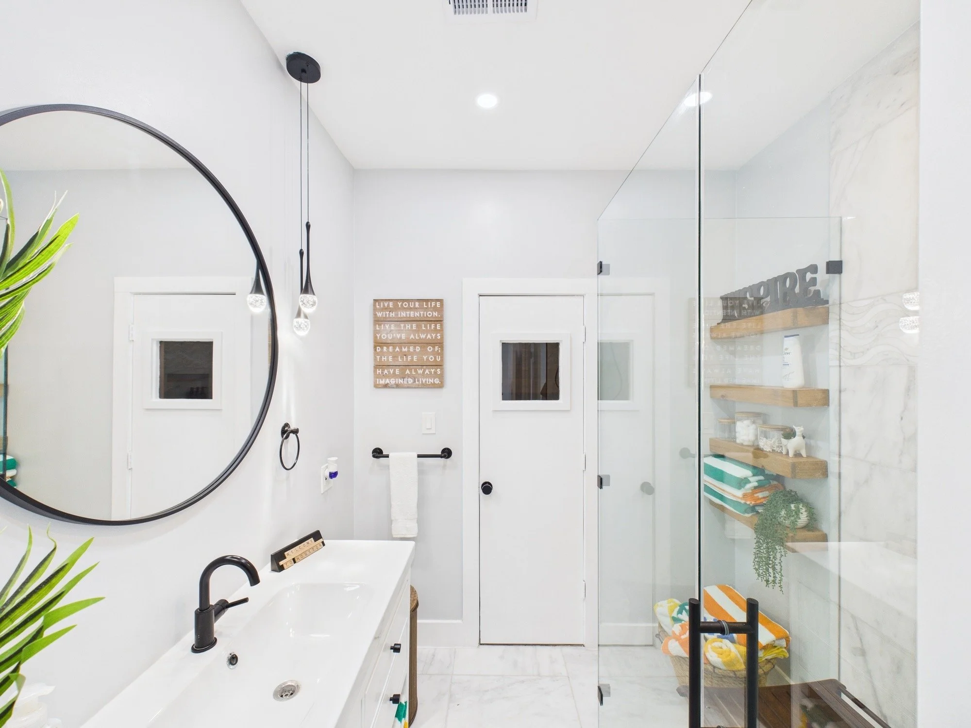 Modern bathroom with a white bathtub, black fixtures, a large round mirror, hanging black pendant lights, a glass shower enclosure, wooden shelves with towels and decor, and framed wall art.