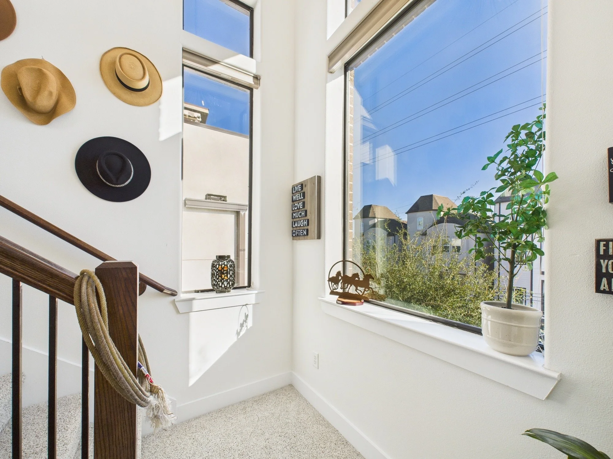 A sunlit window area with a houseplant in a white vase on the windowsill, decorative hats hanging on the wall, a small lantern, and a sign with the words "LIVE WELL LOVE MUCH LAUGH OFTEN". Outside the window, rooftops and power lines are visible agai