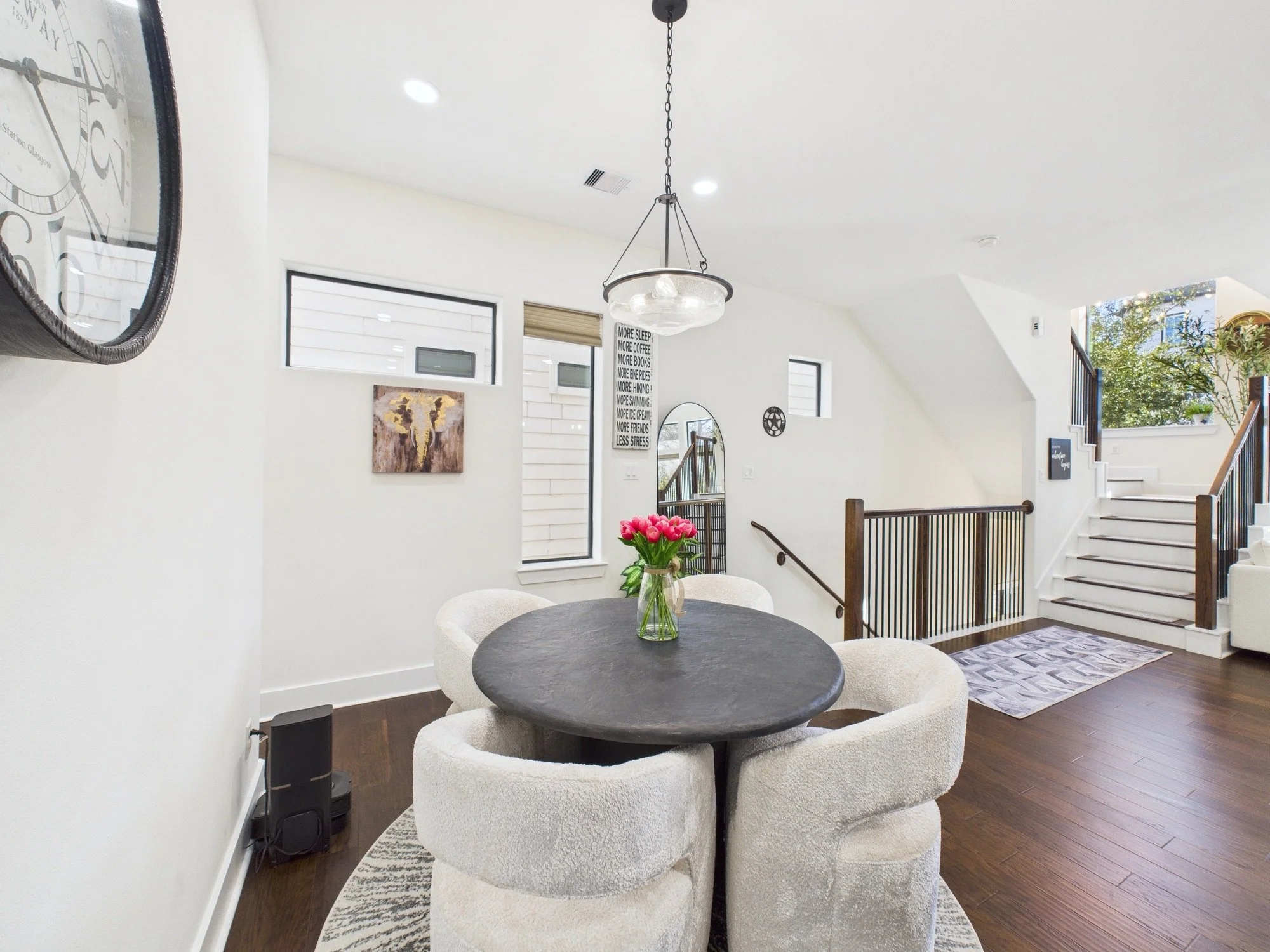 A modern living space with white walls and dark wooden flooring. A round black table with a vase of pink tulips is surrounded by cozy white armchairs. The staircase has wooden steps and black railings, leading to an upper level with window views of t