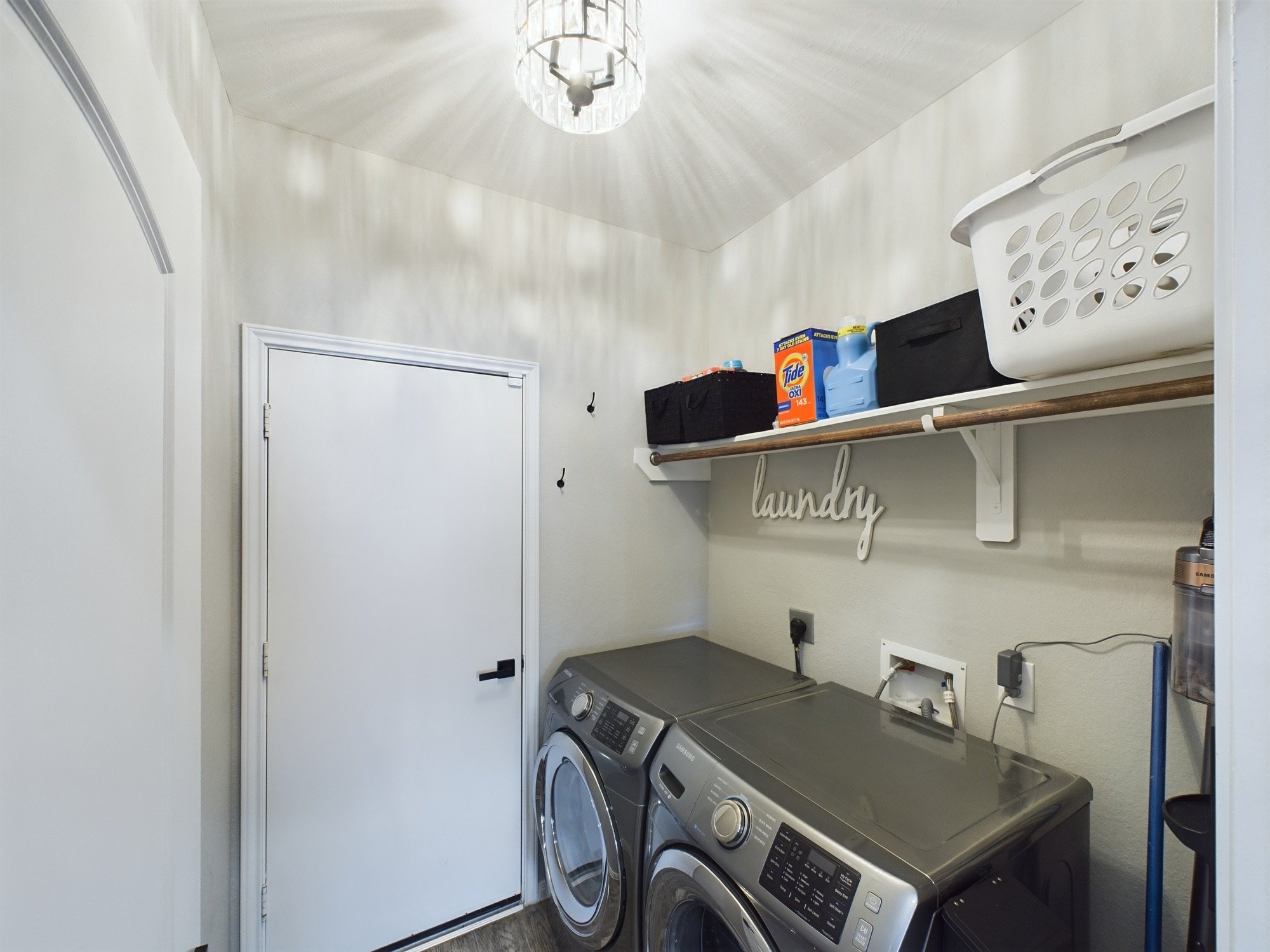 A laundry room with a white door, a shelf holding laundry supplies, a washing machine, and a dryer.