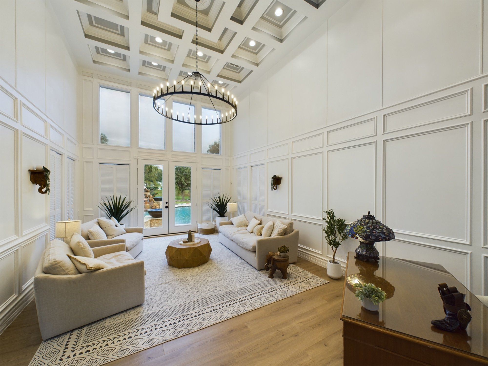 Living room with white panel walls, beige couches, a large window, and a black chandelier with candles hanging from the ceiling.