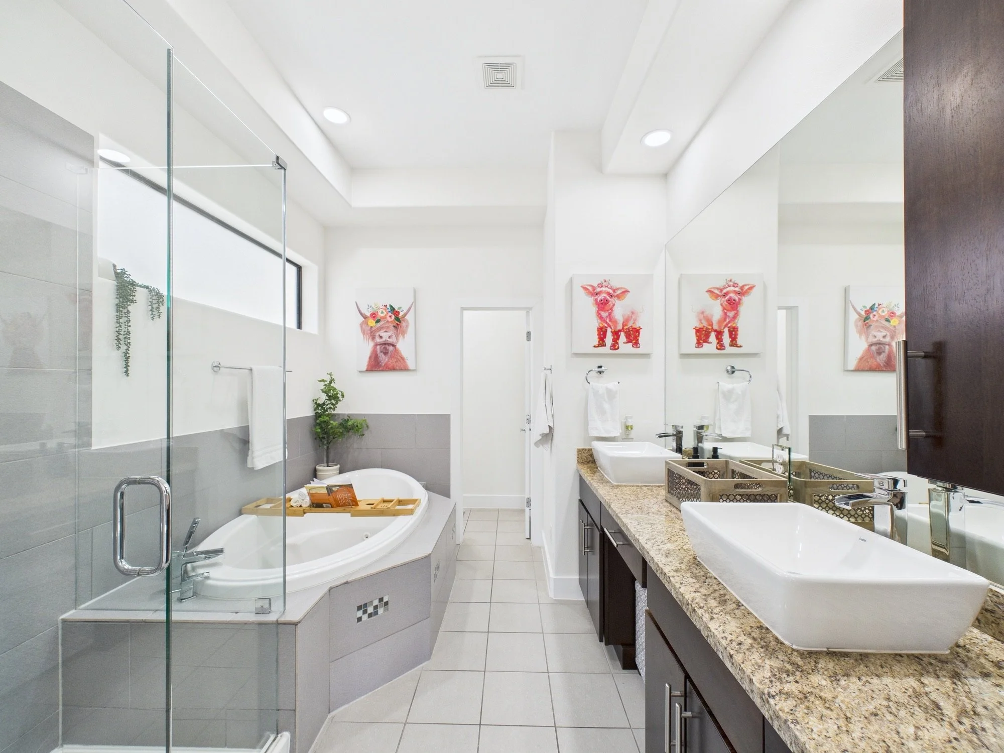 Modern bathroom with white walls, a walk-in shower with glass doors, a bathtub with a wooden tray, a granite countertop with dual sinks, and colorful pig-themed artwork on the walls.