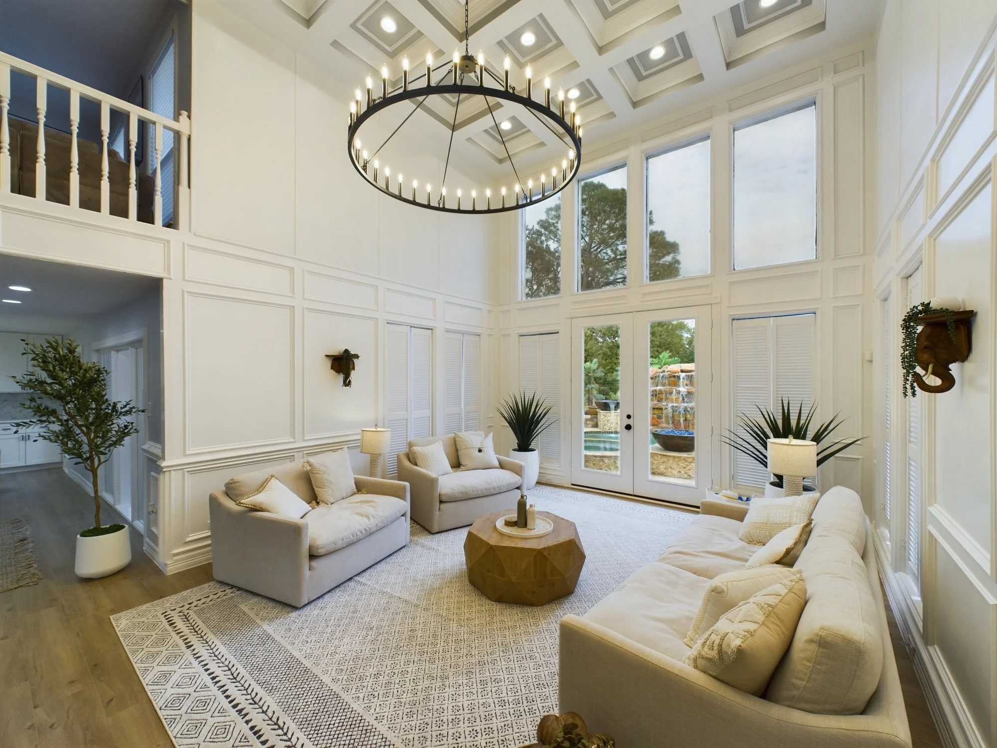 Living room with cream-colored sofas, a geometric wooden coffee table, plants, and large windows with a view of trees and a waterfall outside. Chandelier hanging from ceiling and wall decor.