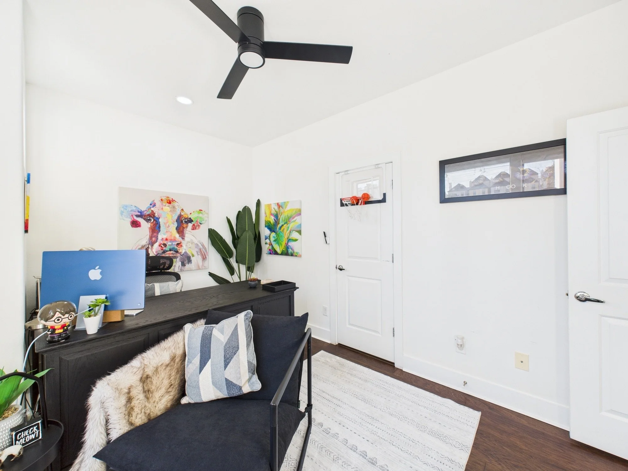 A modern, minimalist living room with white walls and dark wooden floors. There is a black table with a chair, grey pillow, and a furry throw. Artwork and plants are on the wall, and a ceiling fan is above.