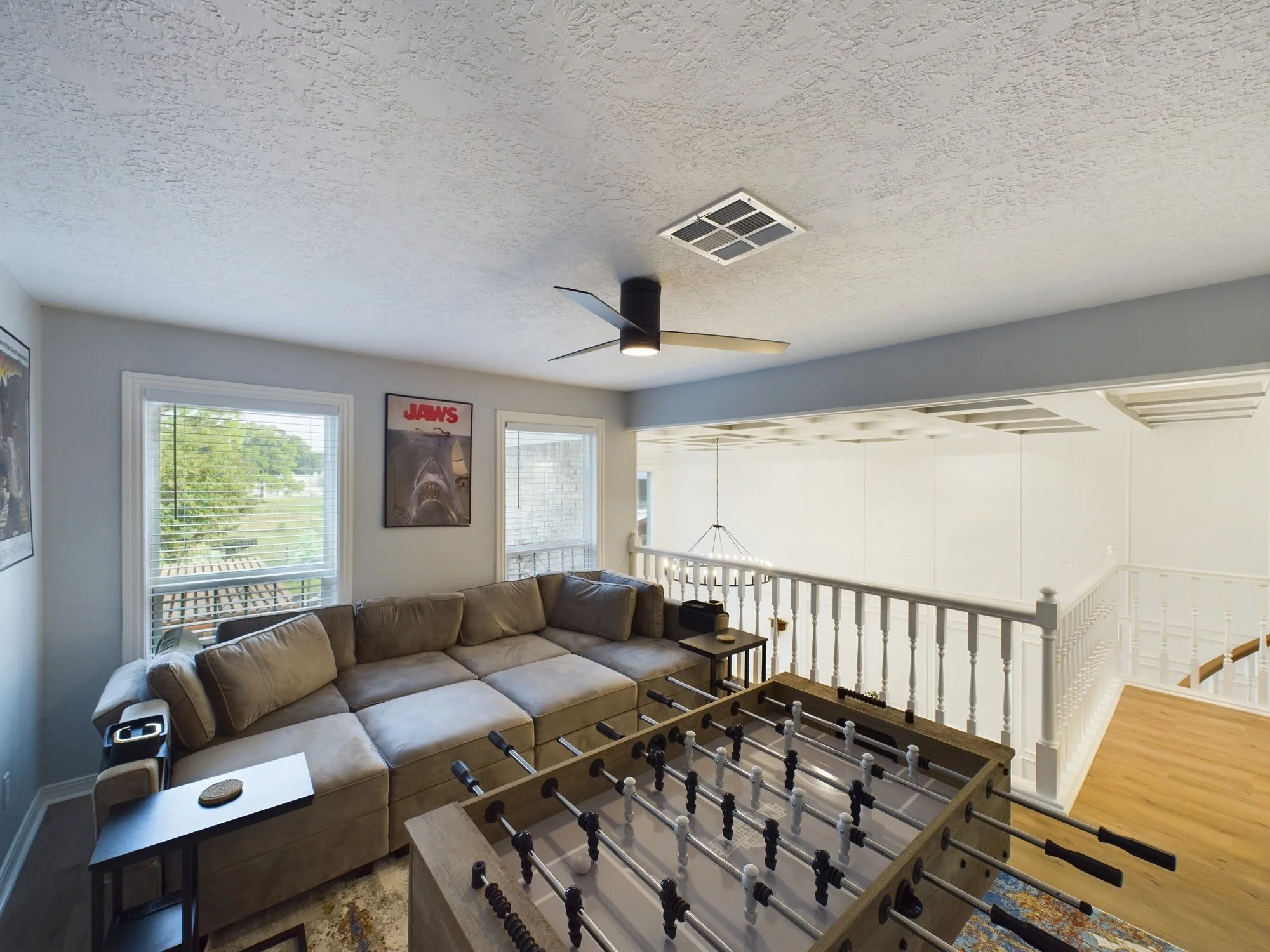 Living room with beige sectional sofa, large windows, wall art, and a foosball table in the center.