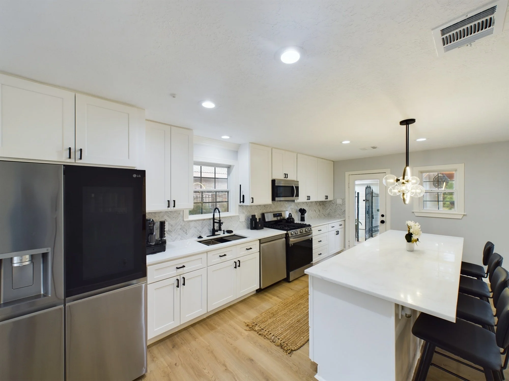 Modern kitchen with white cabinets, stainless steel appliances, and a white island. Pendant light fixture hanging above the island, and a black dining set with chairs. Recessed lighting in the ceiling and windows letting in natural light.