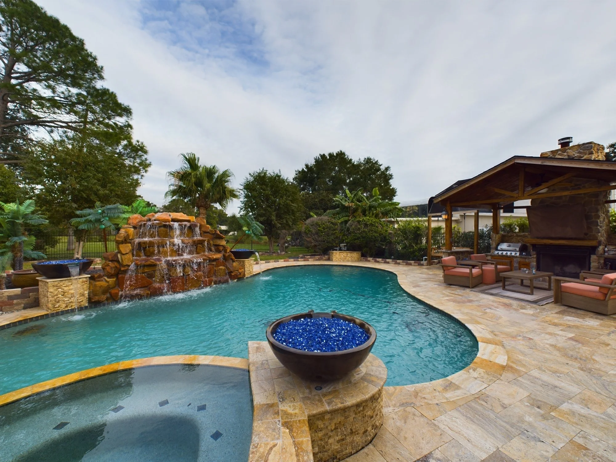 A backyard pool area with a rock waterfall feature, surrounded by tropical plants, a covered seating area, and a clear sky.