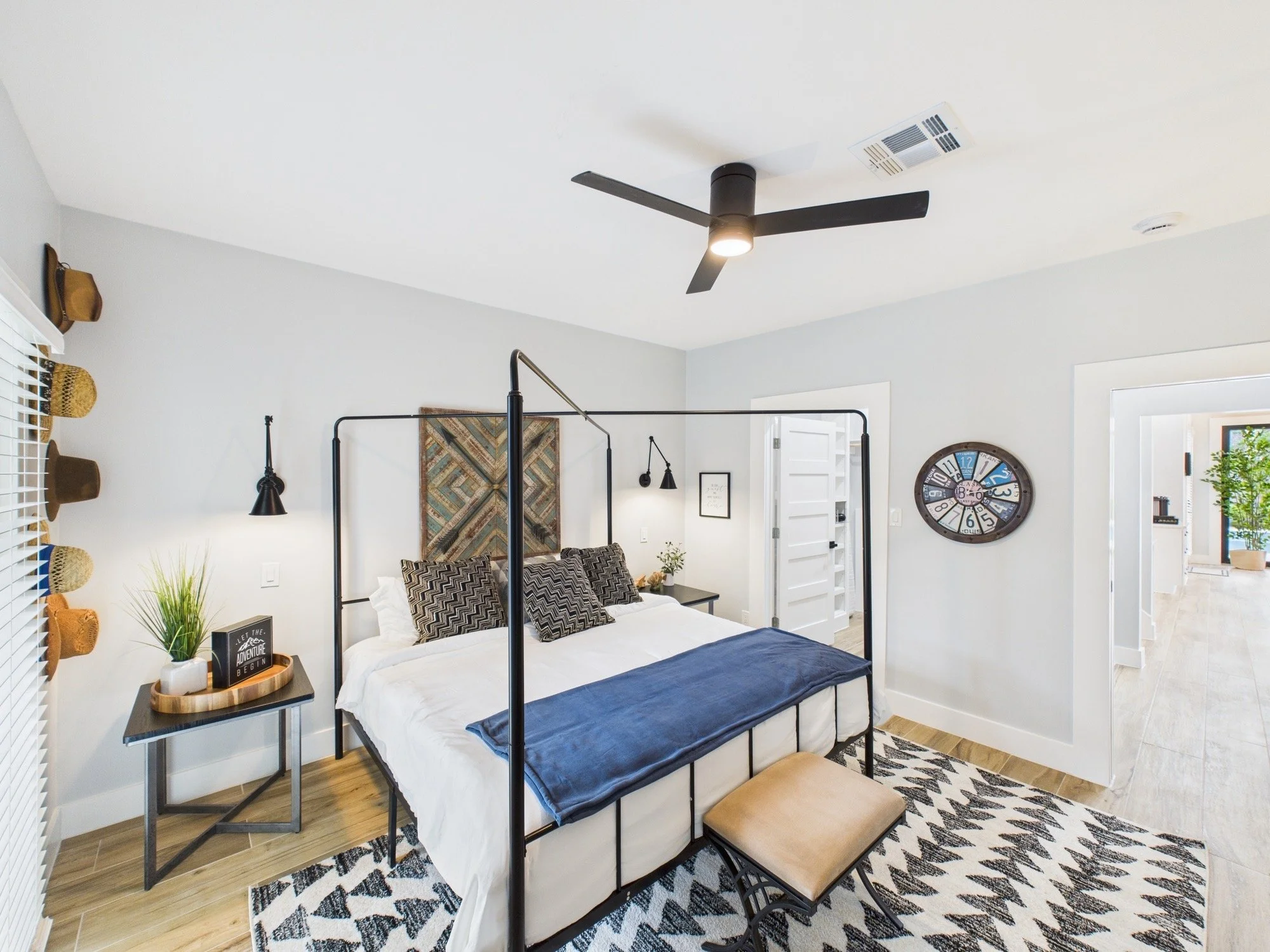A bedroom with a black metal bed frame, white bedding, and decorative pillows. There is a bedside table with a plant and a framed quote. Black pendant lights hang beside the bed. A large wall clock is on the wall, and a patterned rug covers part of t